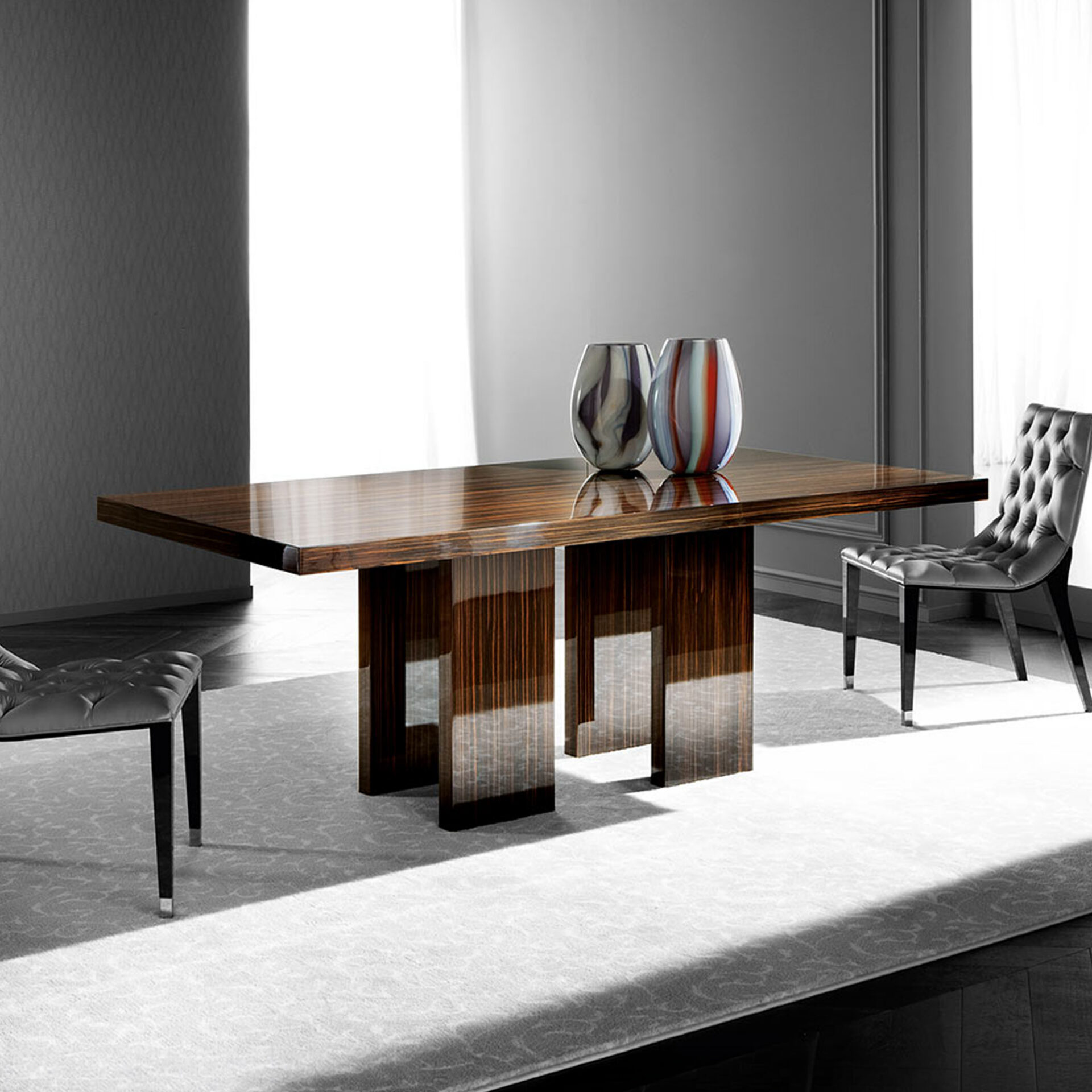 A modern dining room featuring a large, rectangular, highly polished dark wood table with three pedestal legs, two tufted dark chairs, and two colorful decorative vases on the table, all set on a light patterned rug.