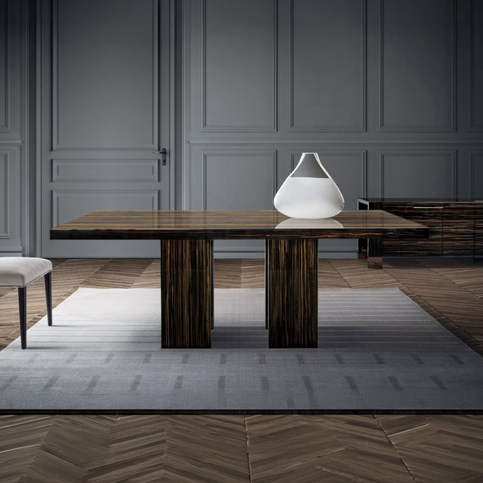 A luxurious dining room with a glossy dark wood table, a white vase, and a light gray rug on a herringbone wood floor, against dark gray paneled walls.