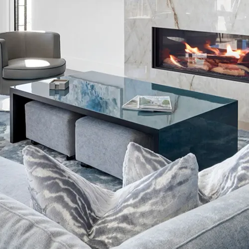 A modern living room with a gray sofa, a dark reflective coffee table with ottomans, a gray armchair, and a marble fireplace with flames.