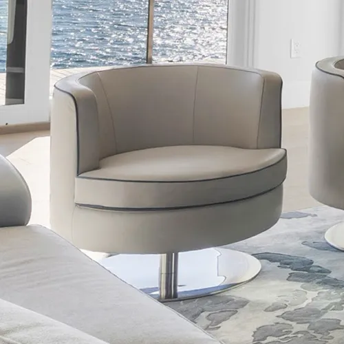 A light beige barrel swivel chair with dark piping on a chrome base, positioned in front of a window overlooking sparkling water.