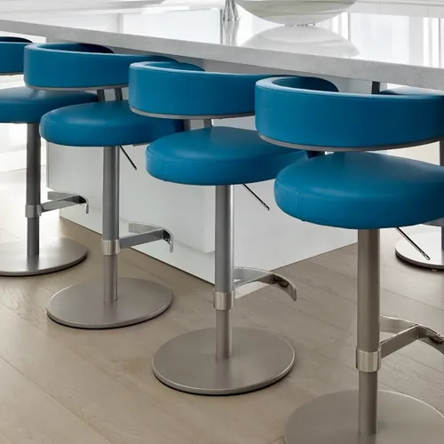Five blue bar stools with brushed metal bases lined up against a light-colored kitchen counter on a light wood floor.