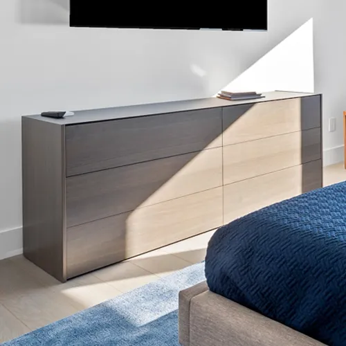 A modern gray wooden dresser with six drawers, a black TV above it, and a bed with a blue comforter in a sunlit room.
