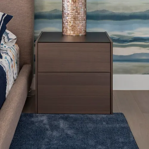 A dark brown nightstand with a textured copper lamp next to a bed with blue bedding and a blue rug on a wooden floor, against a blue and white abstract wall.