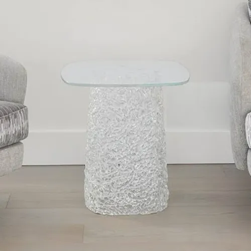 A clear glass side table with a textured, cylindrical base stands between two light gray armchairs on a light wood floor.