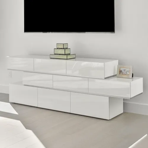 A white, glossy, modular media console with a wall-mounted black TV above it. Decorative green glass boxes and a framed picture are on the console. Light-colored floor with shadows.