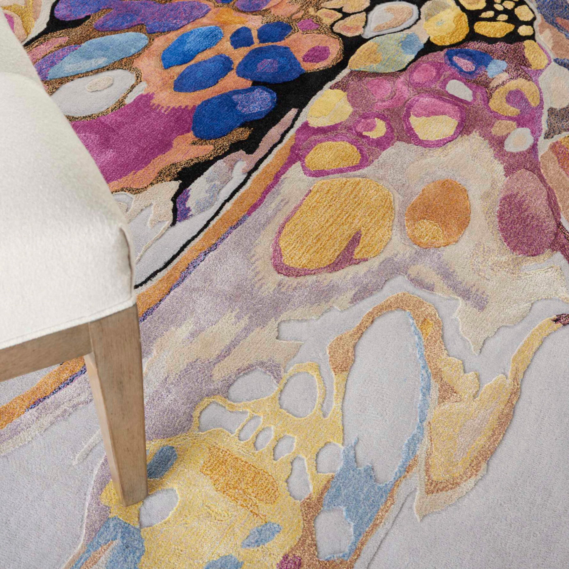 Close-up of a vibrant, abstract rug with organic shapes in blue, fuchsia, yellow, and orange, featuring varying pile heights. A cream upholstered chair with a light wooden leg is partially visible on the left.