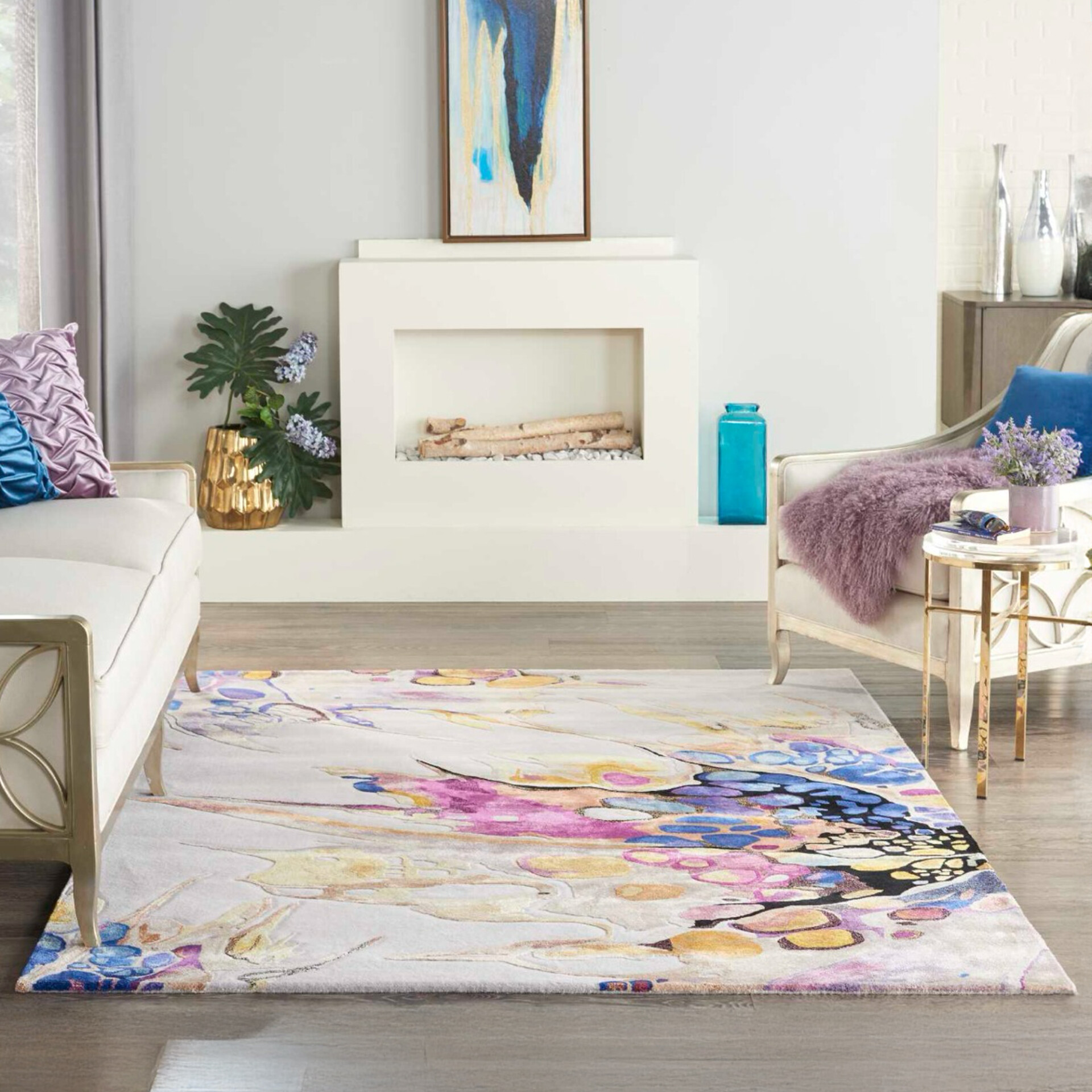 A modern living room featuring a large, colorful abstract floral rug on a light wood floor. The room includes white and gold-framed furniture, a minimalist white fireplace, and various decorative accents in blue, purple, and gold.
