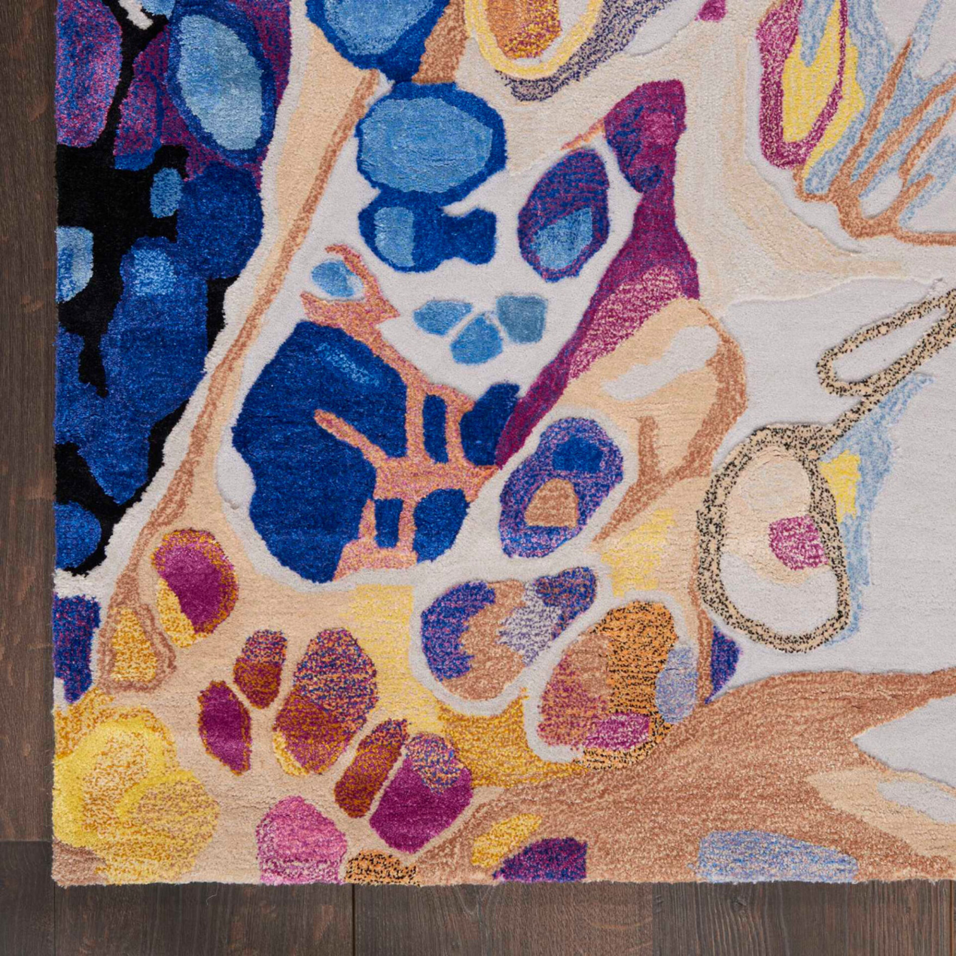 Close-up of a plush, abstract rug with organic shapes in vibrant blues, purples, yellows, and oranges on a cream background, resting on dark wood floorboards.