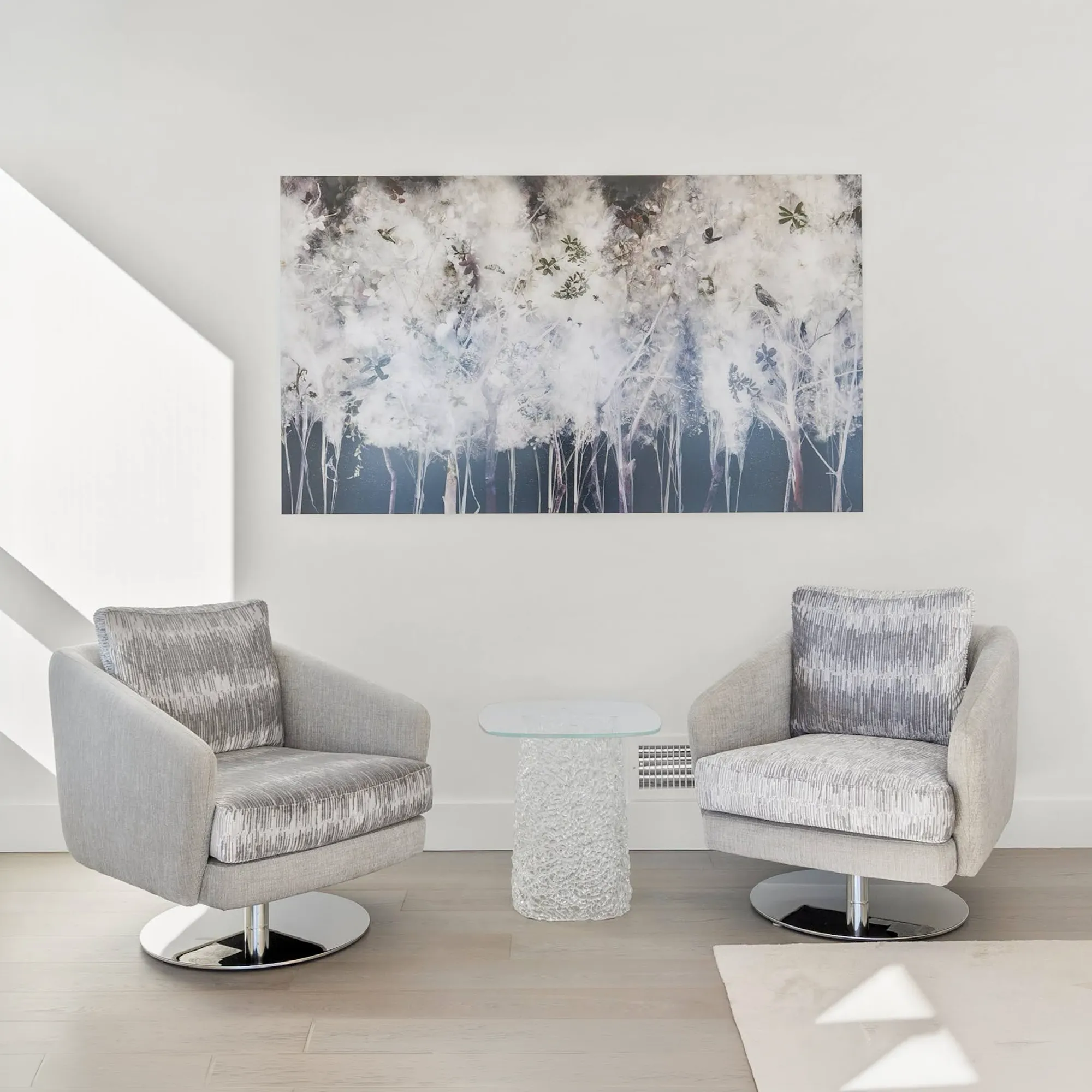 Two light grey swivel armchairs with patterned cushions flank a clear glass side table, beneath a large abstract painting of white fluffy plants and birds on a white wall.