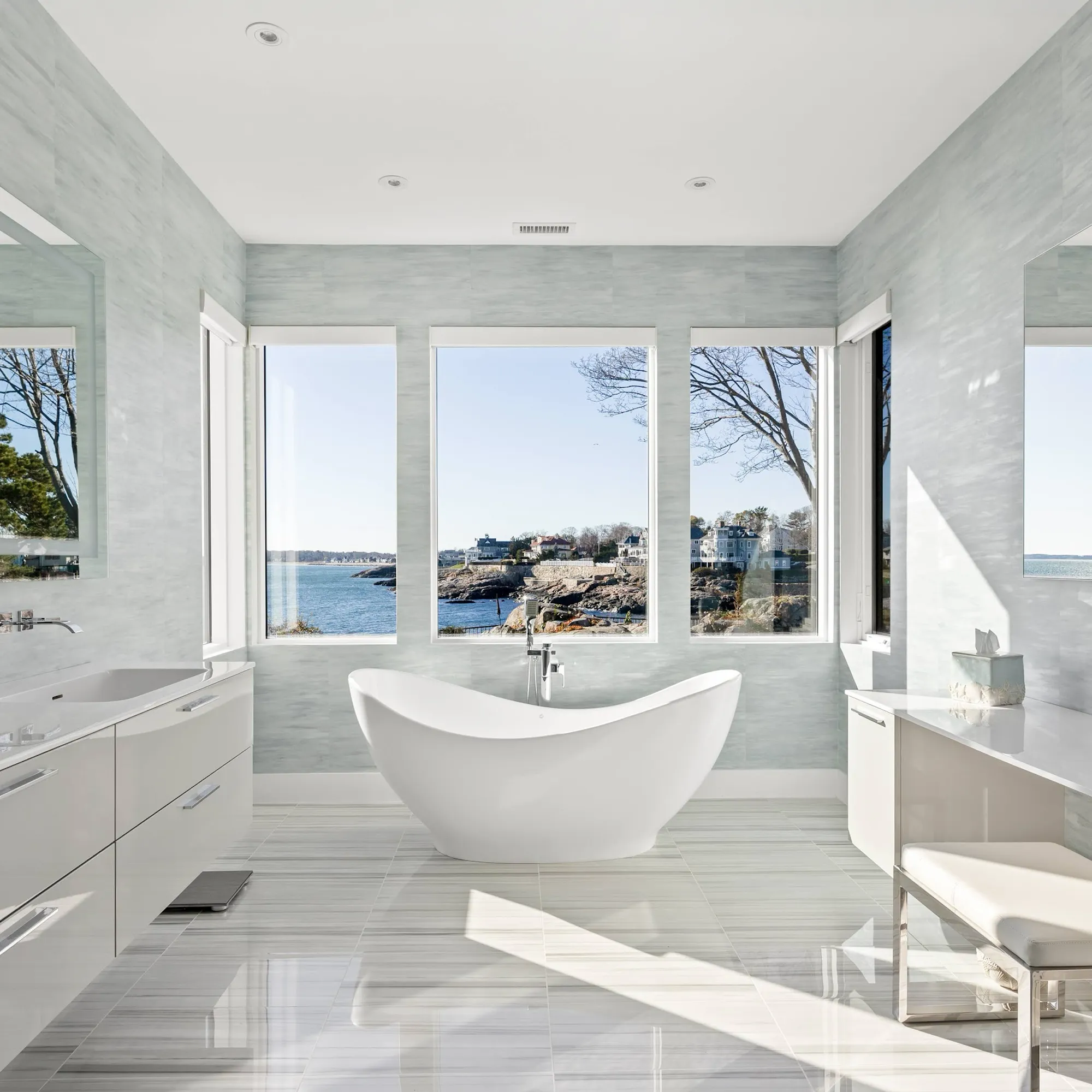A modern bathroom with a white freestanding tub facing large windows with an ocean and coastal town view. White vanities are on either side, and the room has light blue-grey tiled walls and a glossy striped floor.