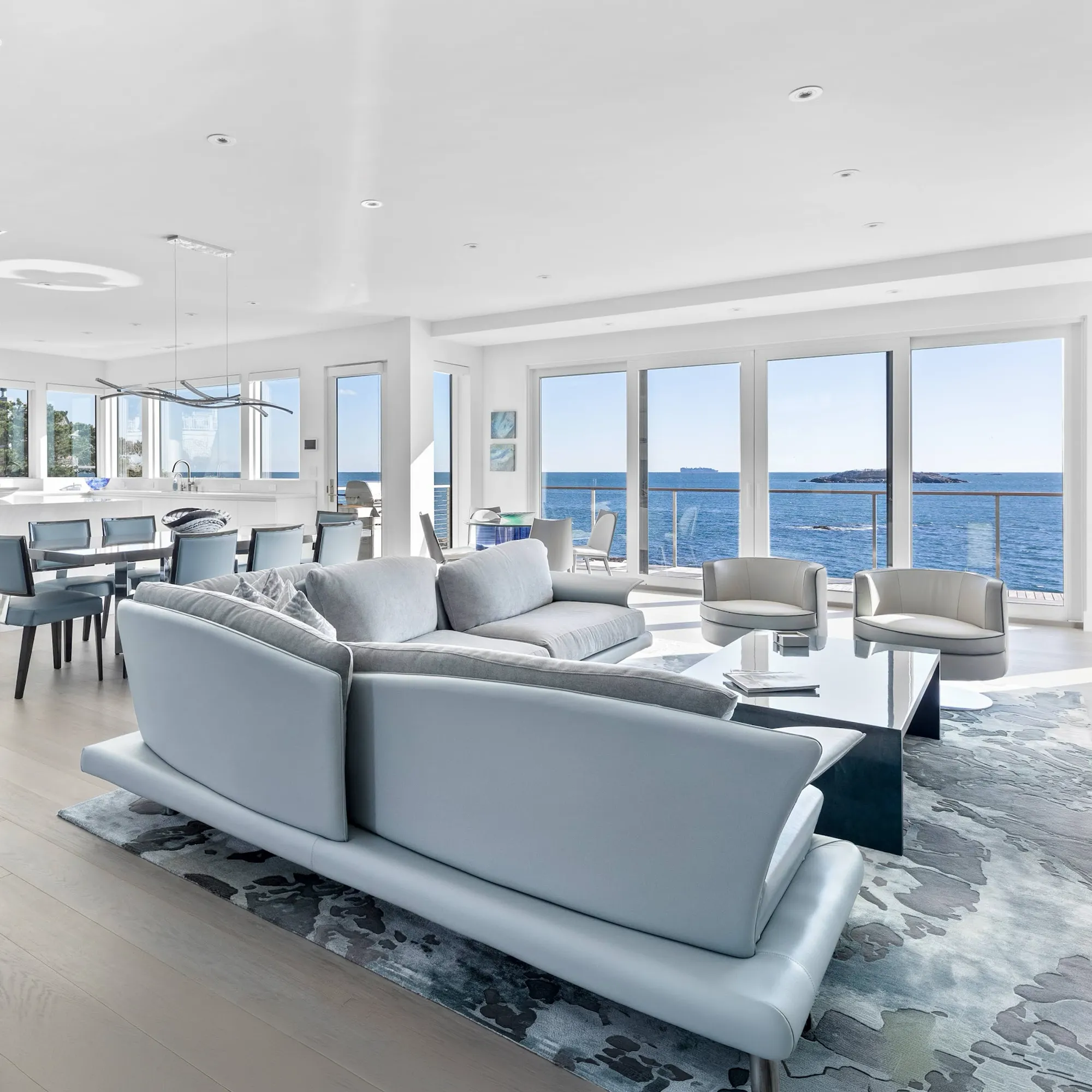 A modern open-concept living room and dining area with light blue and grey furniture, featuring floor-to-ceiling windows offering a panoramic view of the blue ocean with small islands and a ship on the horizon.