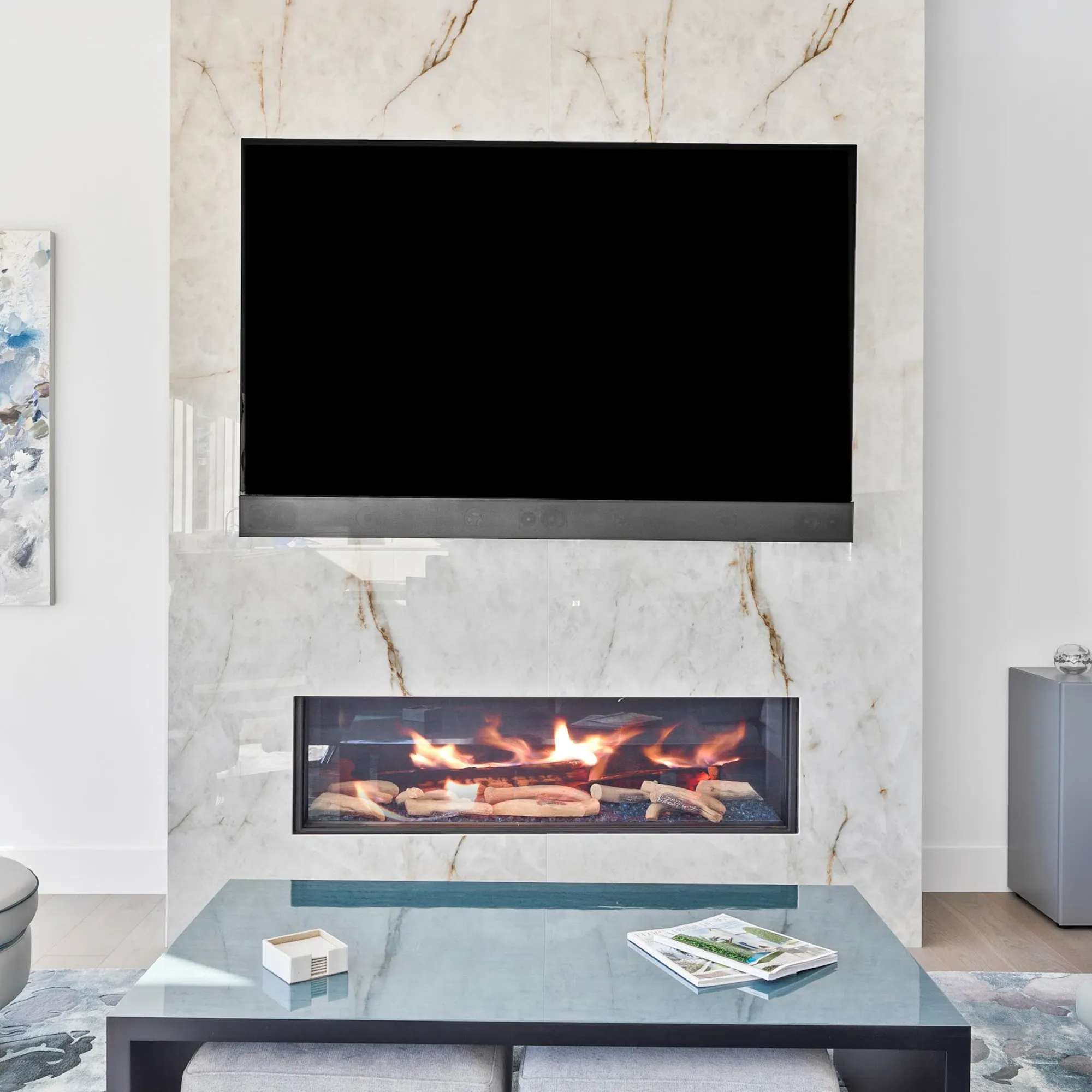 A modern living room with a large TV above a linear gas fireplace, both set into a white marble wall. A glass coffee table sits in front.