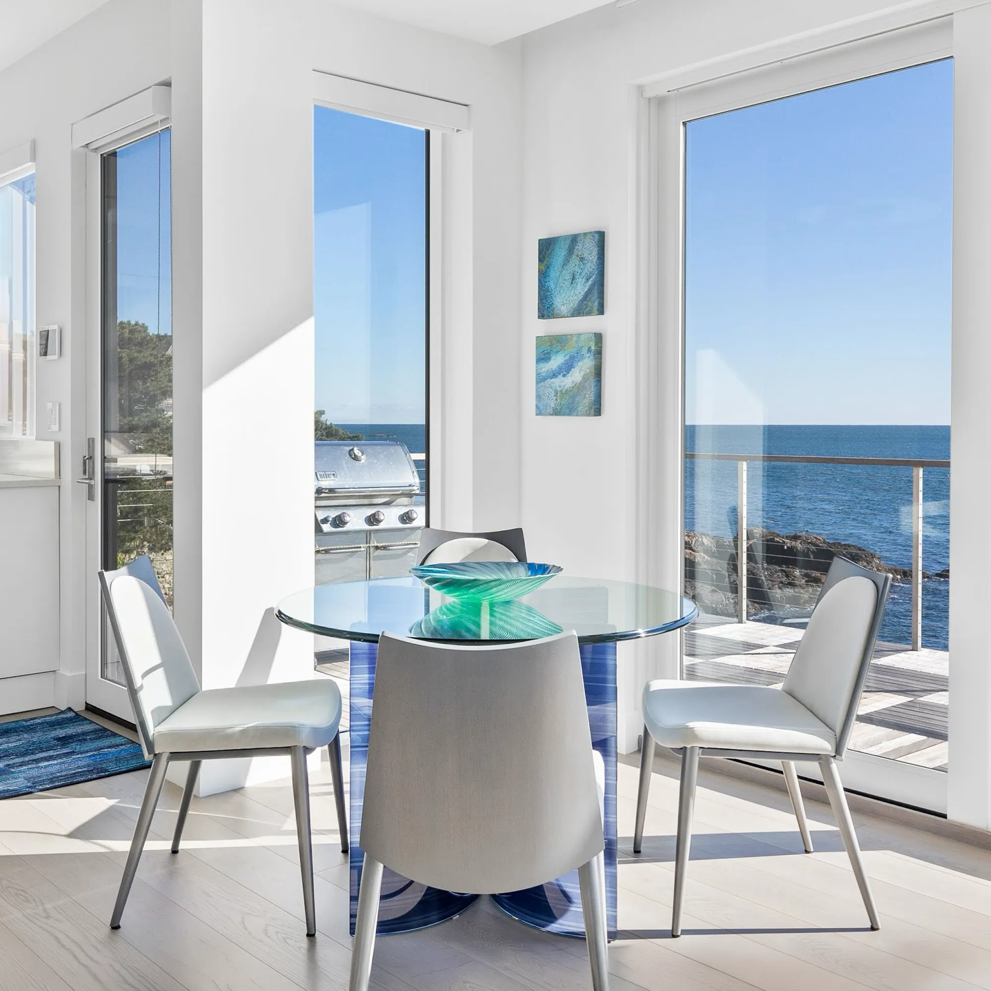 A modern dining area with a round glass table and four light gray chairs, overlooking a vast blue ocean and rocky coastline through large windows.