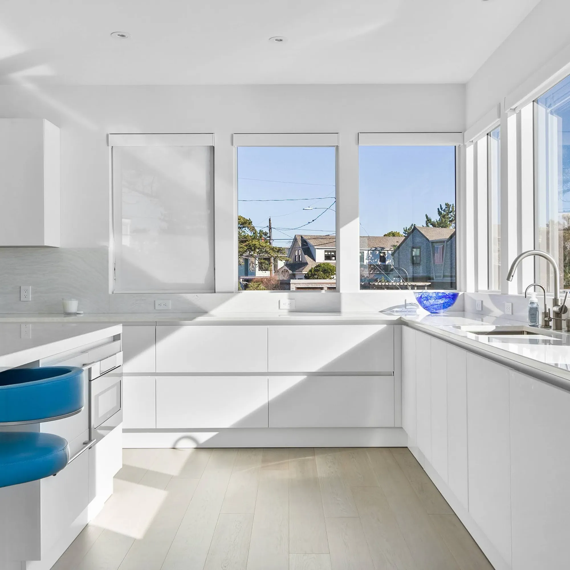 A modern white kitchen with an L-shaped counter, light wood floors, and large windows overlooking a residential neighborhood. Blue bar stools and a blue bowl add color.