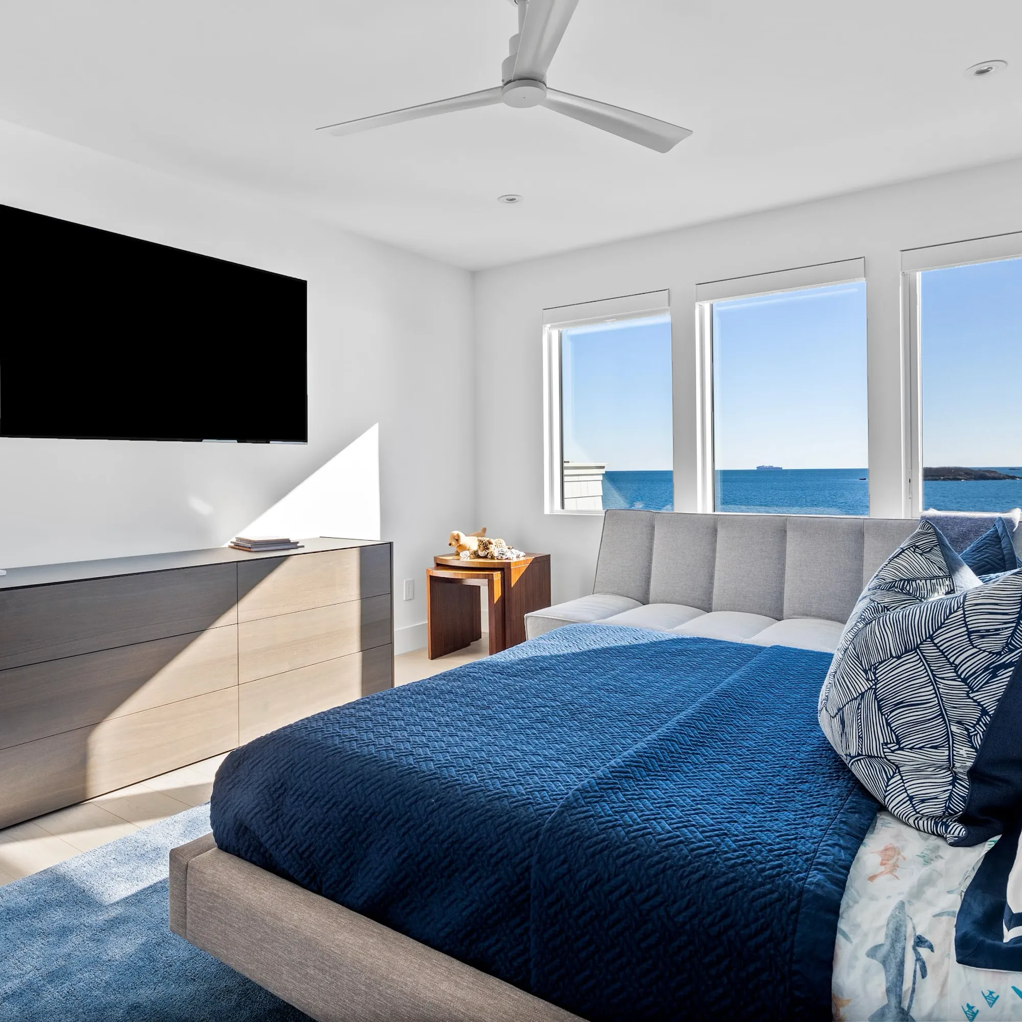 A modern bedroom with a blue bed, dark wood dresser, and large windows offering a clear view of the blue ocean.