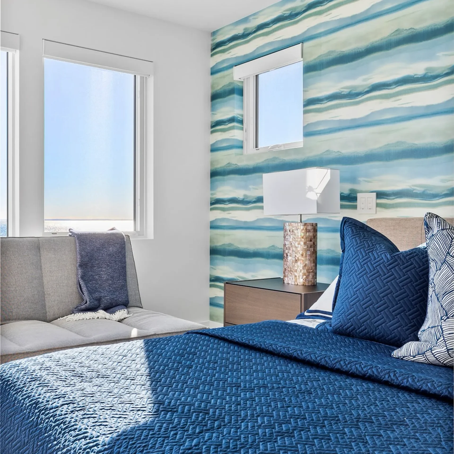 A modern bedroom with a blue quilted bed, gray sofa, and windows overlooking the ocean. An accent wall features blue and green wavy wallpaper.