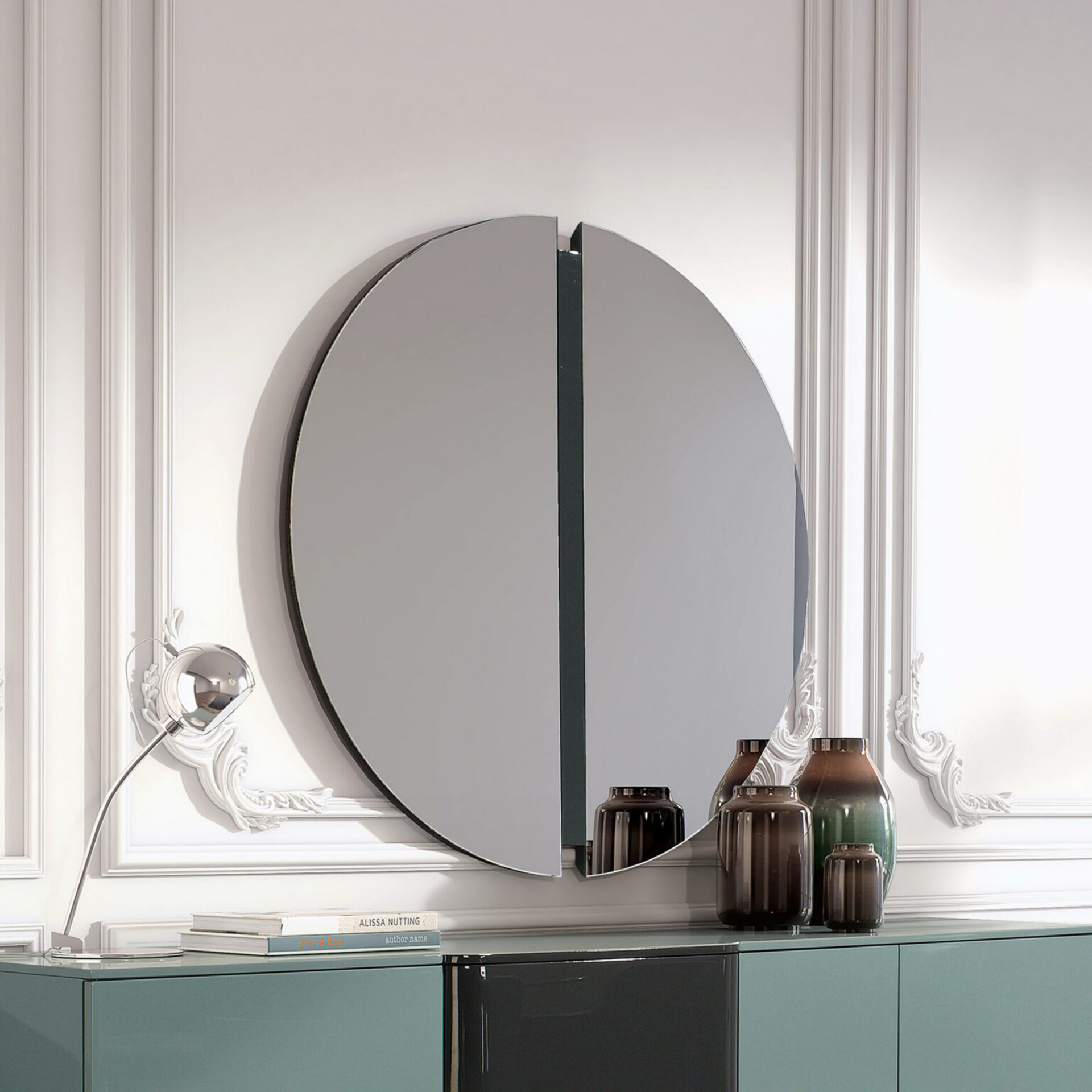 A modern split-circle mirror hangs above a teal and dark gray sideboard. On the sideboard are a silver lamp, two books, and three glass vases. The wall behind is white with classic decorative paneling.