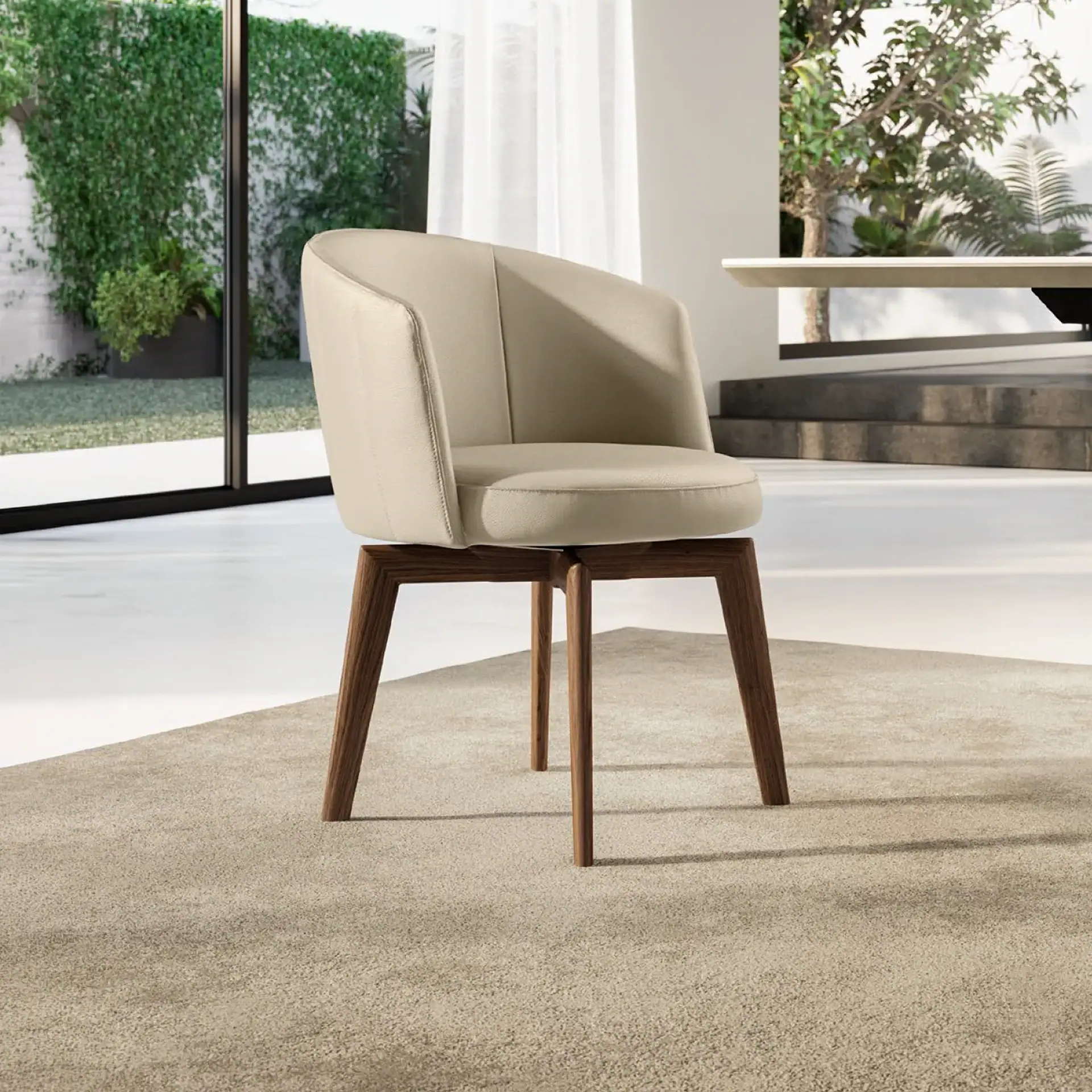 A modern beige upholstered chair with dark brown wooden legs sits on a light beige rug in a bright room with large windows overlooking lush green foliage.