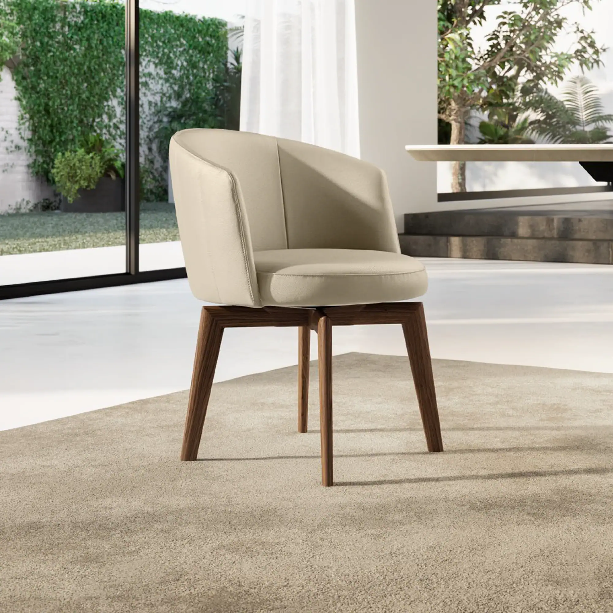 A modern beige upholstered chair with dark brown wooden legs sits on a light beige rug in a bright room with large windows overlooking lush green foliage.