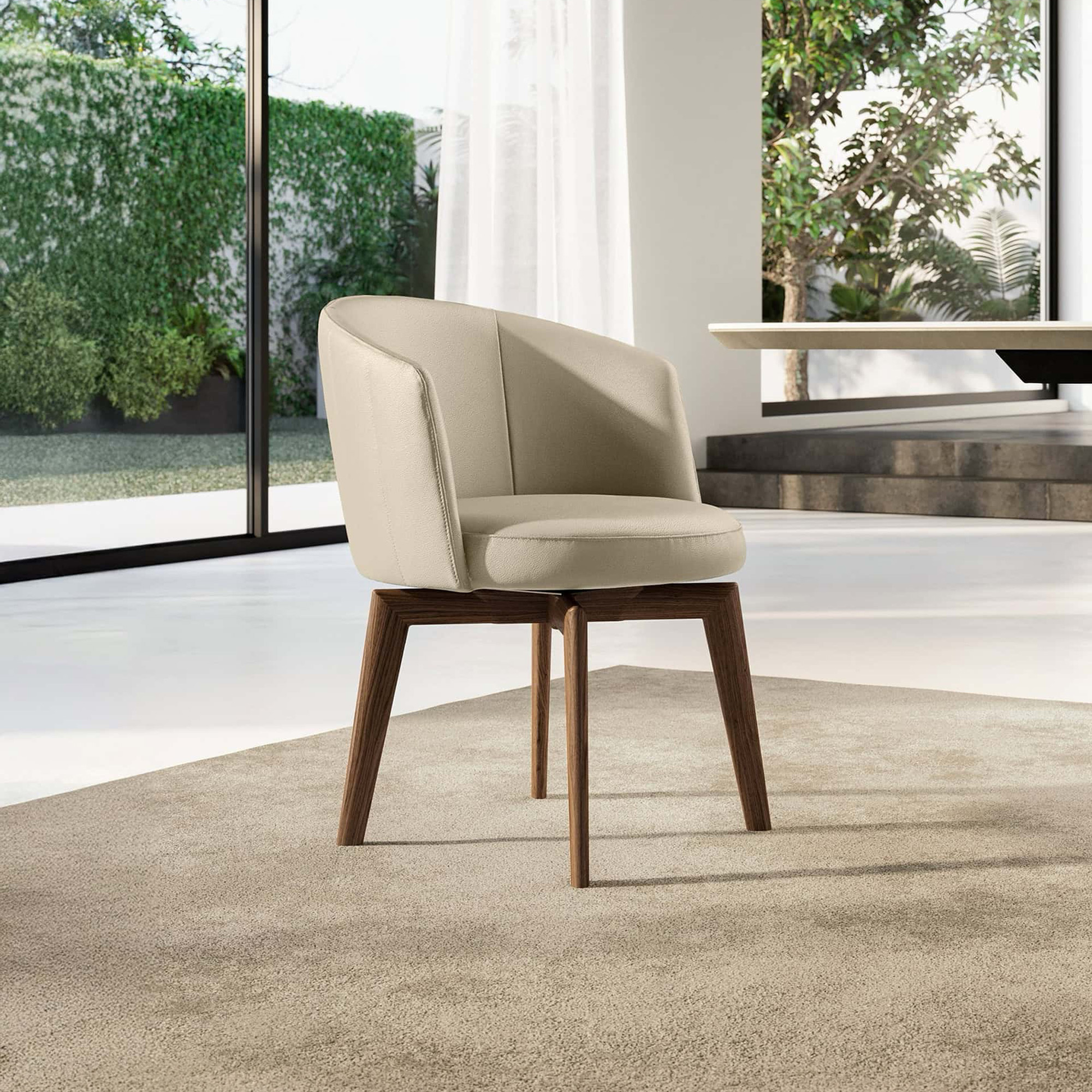 A modern beige upholstered chair with dark brown wooden legs sits on a light beige rug in a bright room with large windows overlooking lush green foliage.