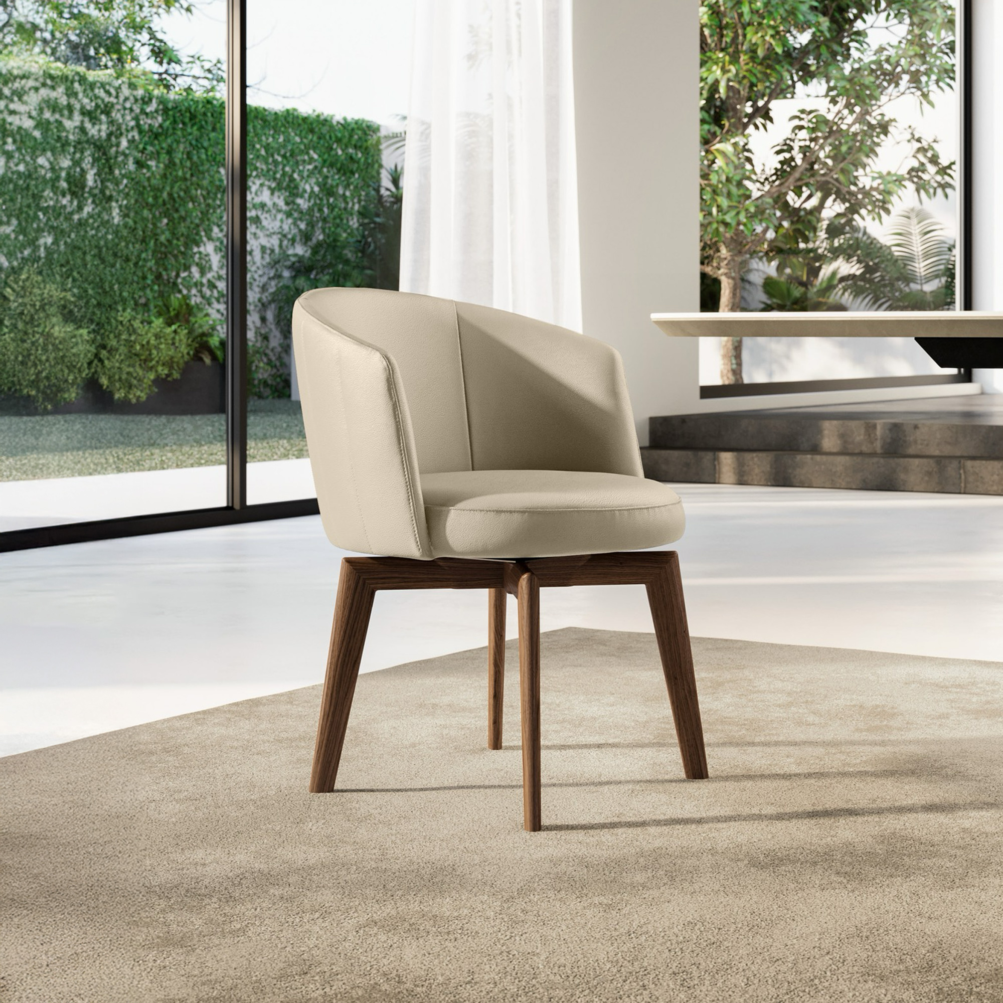 A modern beige upholstered chair with dark brown wooden legs sits on a light beige rug in a bright room with large windows overlooking lush green foliage.