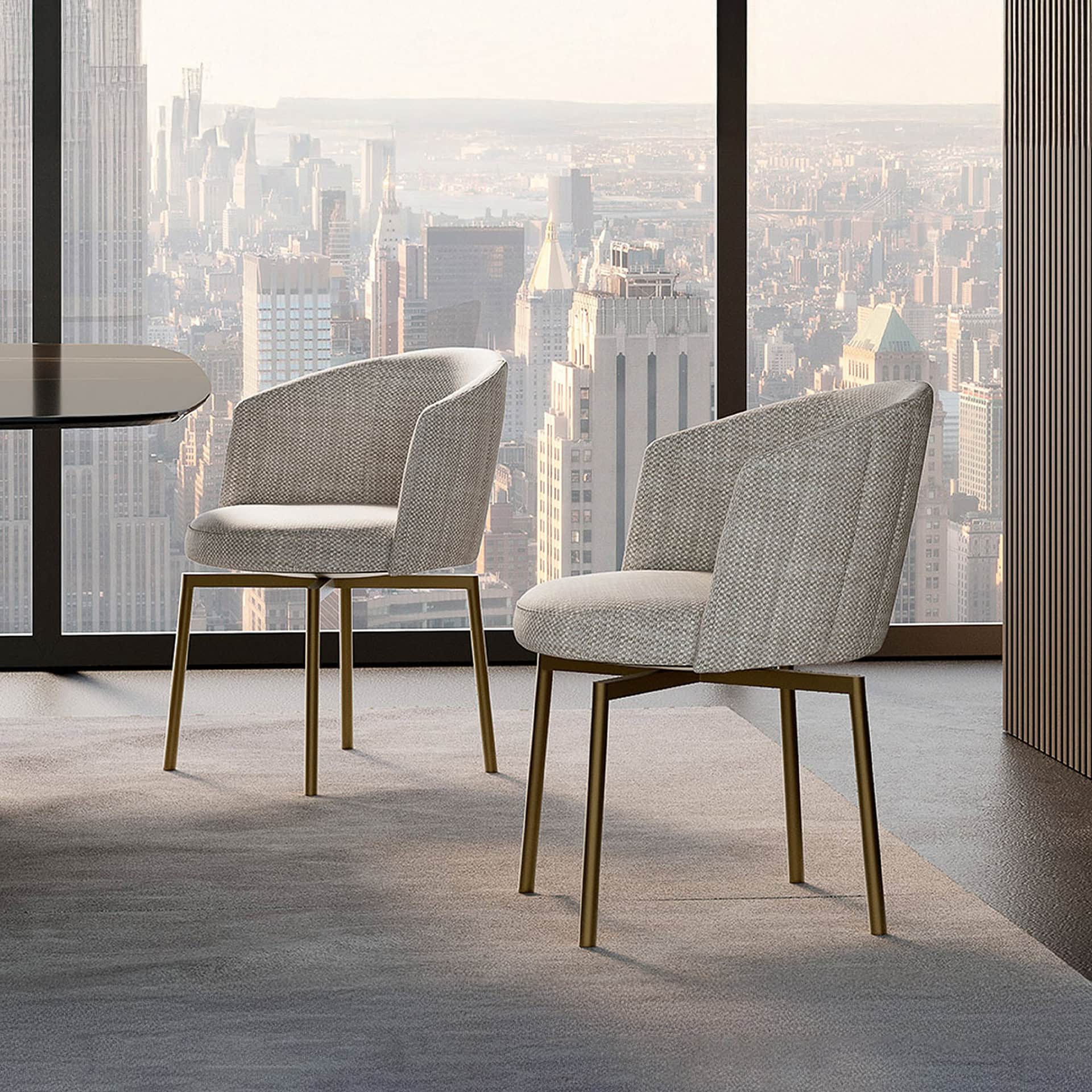 Two modern light gray upholstered chairs with golden legs sit on a light gray rug in front of a large window overlooking a hazy city skyline. A dark table is partially visible on the left.