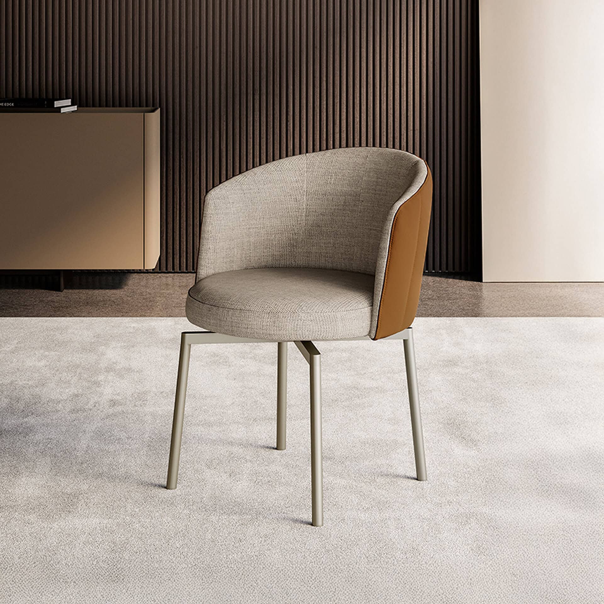 A modern armchair with light beige fabric and tan leather upholstery, featuring curved arms and slender metallic legs, sits on a light carpet. Behind it is a dark brown ribbed wall panel and a matte bronze cabinet.