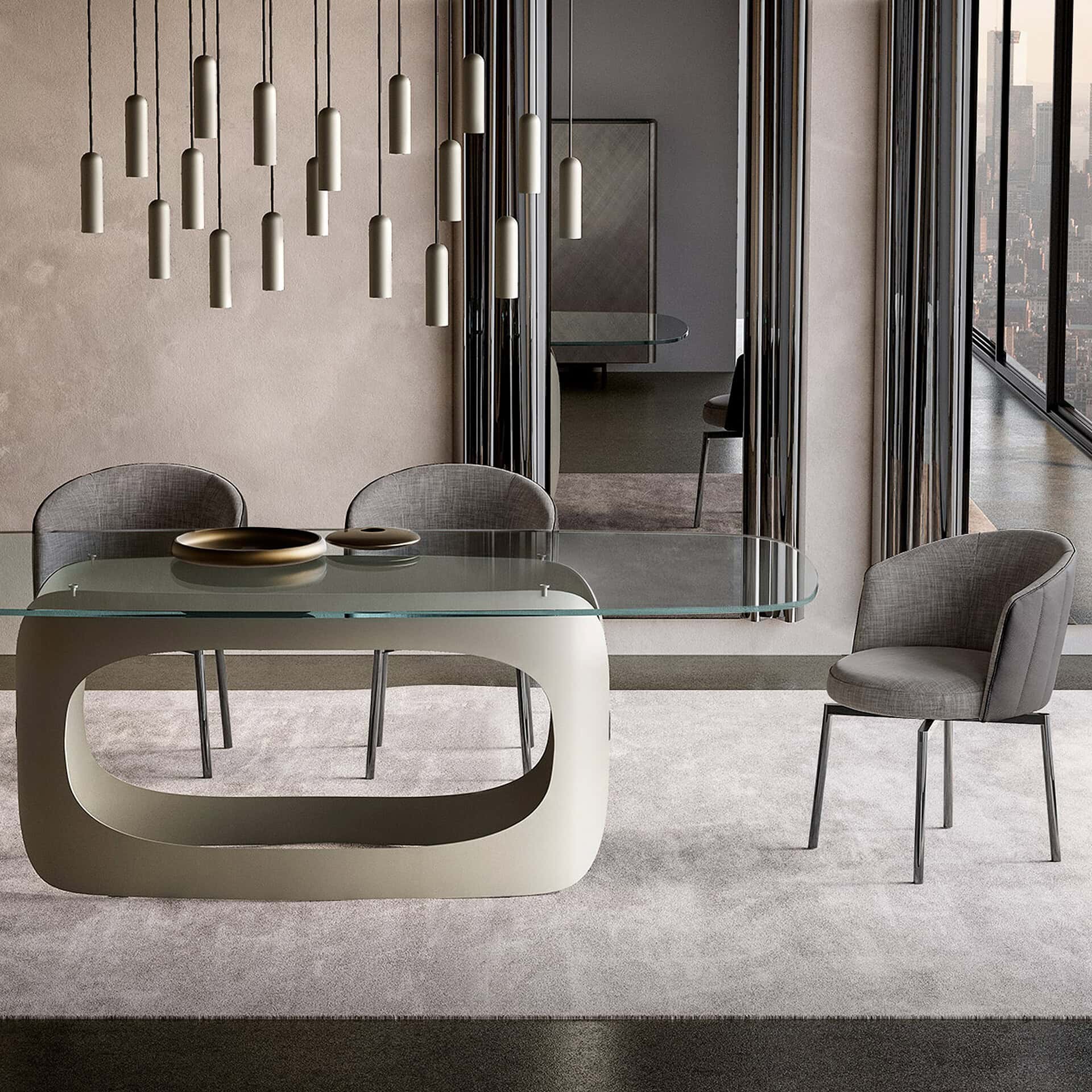 A modern dining room with a glass-top table on a sculptural beige base, surrounded by gray upholstered chairs. Sleek pendant lights hang overhead, and a large window reveals a city skyline. The room has a neutral color palette and plush gray carpet.