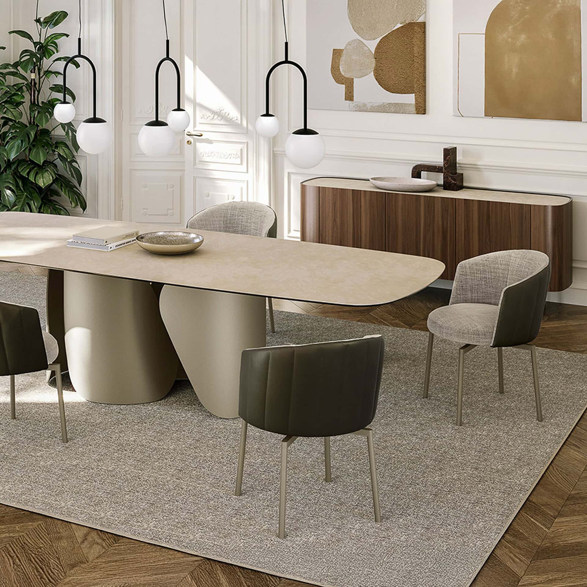 A modern dining room with a large oval beige table, two black U-shaped pendant lights with white globes, and stylish grey and brown chairs on a textured rug. A dark wood credenza and abstract art are in the background.