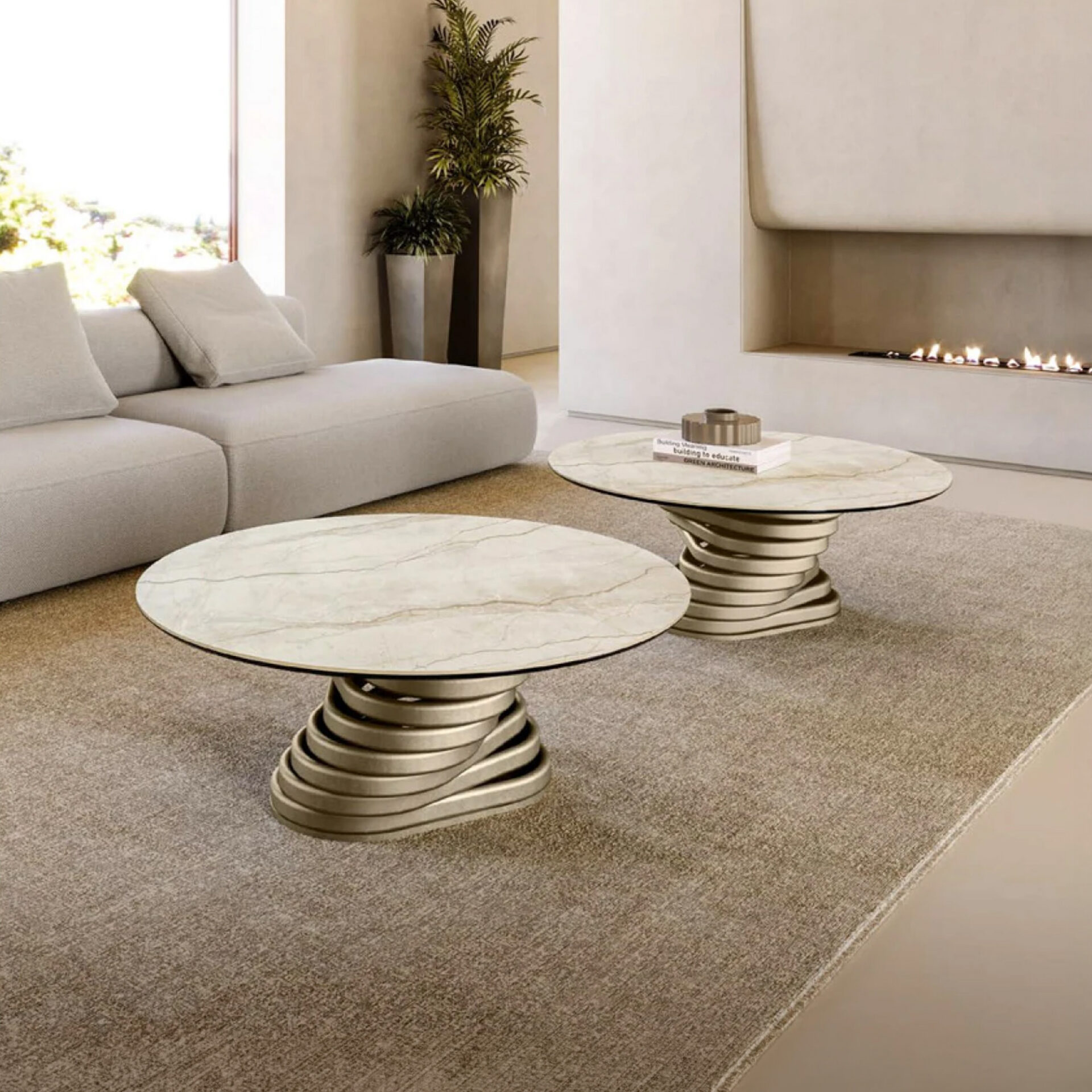Two round marble-top coffee tables with sculptural metallic bases sit on a textured rug in a modern living room. A light grey sofa is to the left, and a linear fireplace with flames is visible in the background.