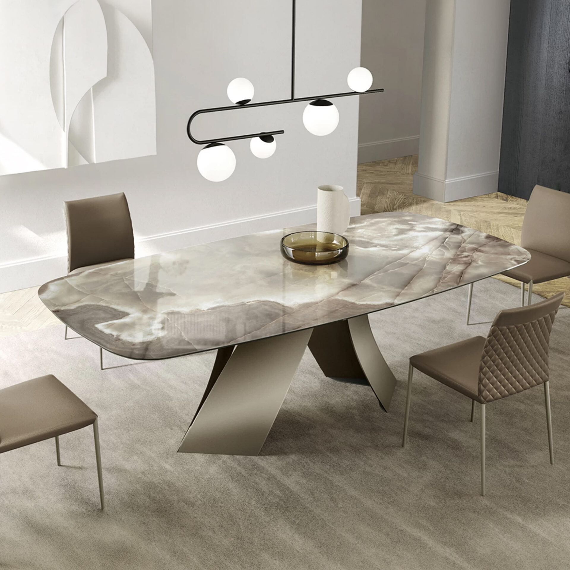 A modern dining room featuring a rectangular natural stone table with a sculptural base, surrounded by taupe upholstered chairs. A black linear chandelier with five spherical lights hangs above the table. A light grey rug covers the floor, and abstract white art adorns the wall.