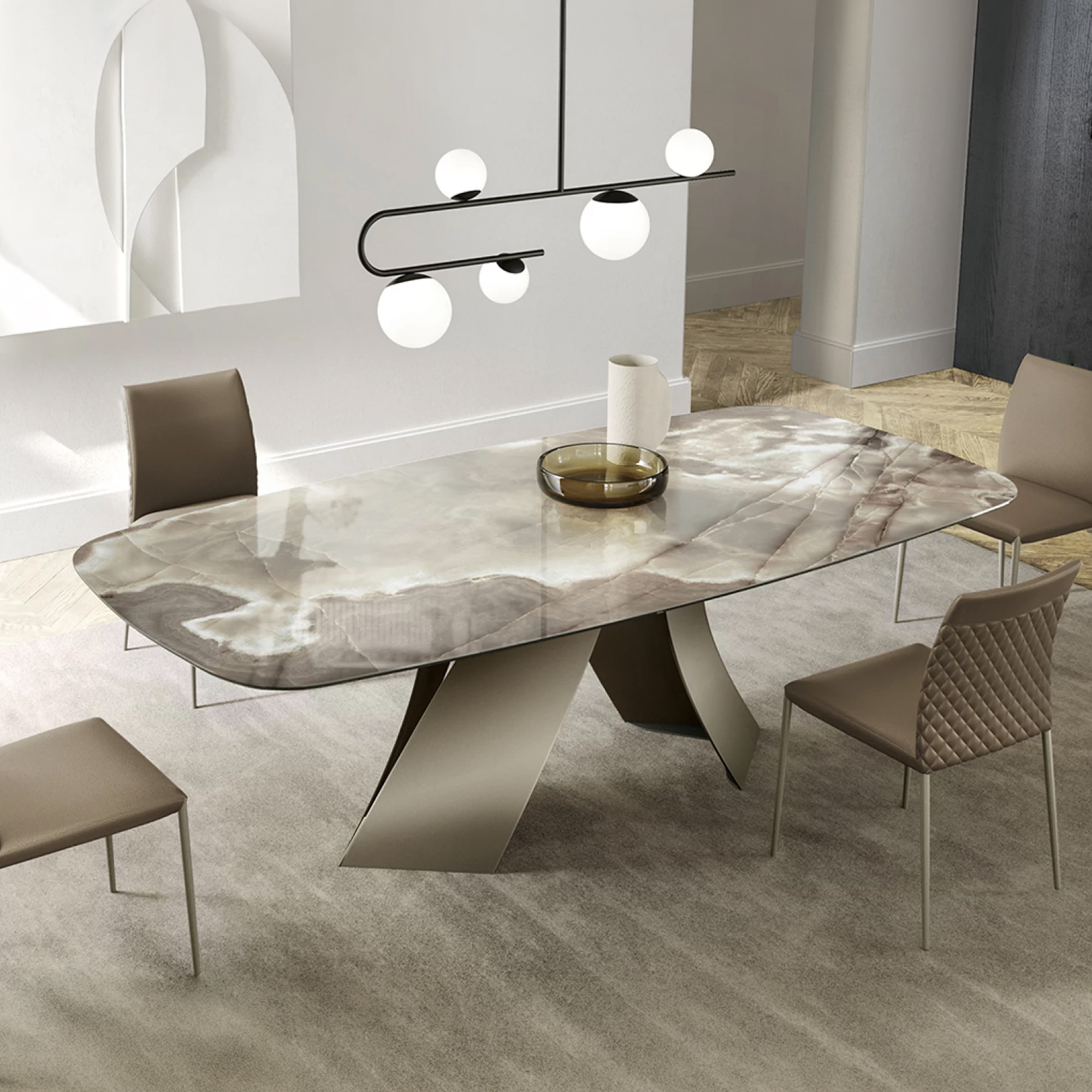 A modern dining room featuring a rectangular natural stone table with a sculptural base, surrounded by taupe upholstered chairs. A black linear chandelier with five spherical lights hangs above the table. A light grey rug covers the floor, and abstract white art adorns the wall.