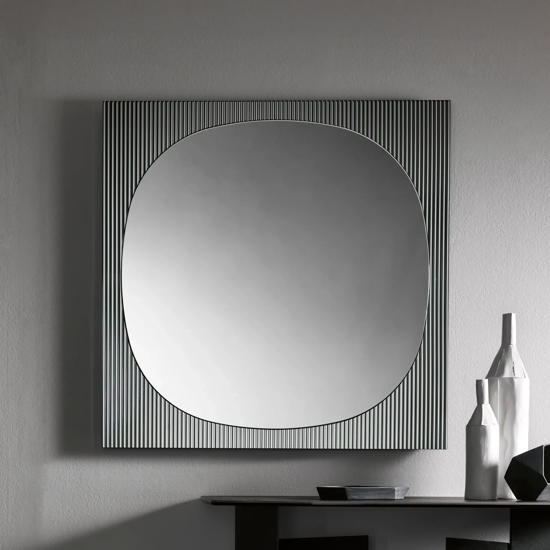 A square mirror with a central oval reflection and a ribbed, vertical-line frame hangs on a gray wall above a dark console table with decorative objects.