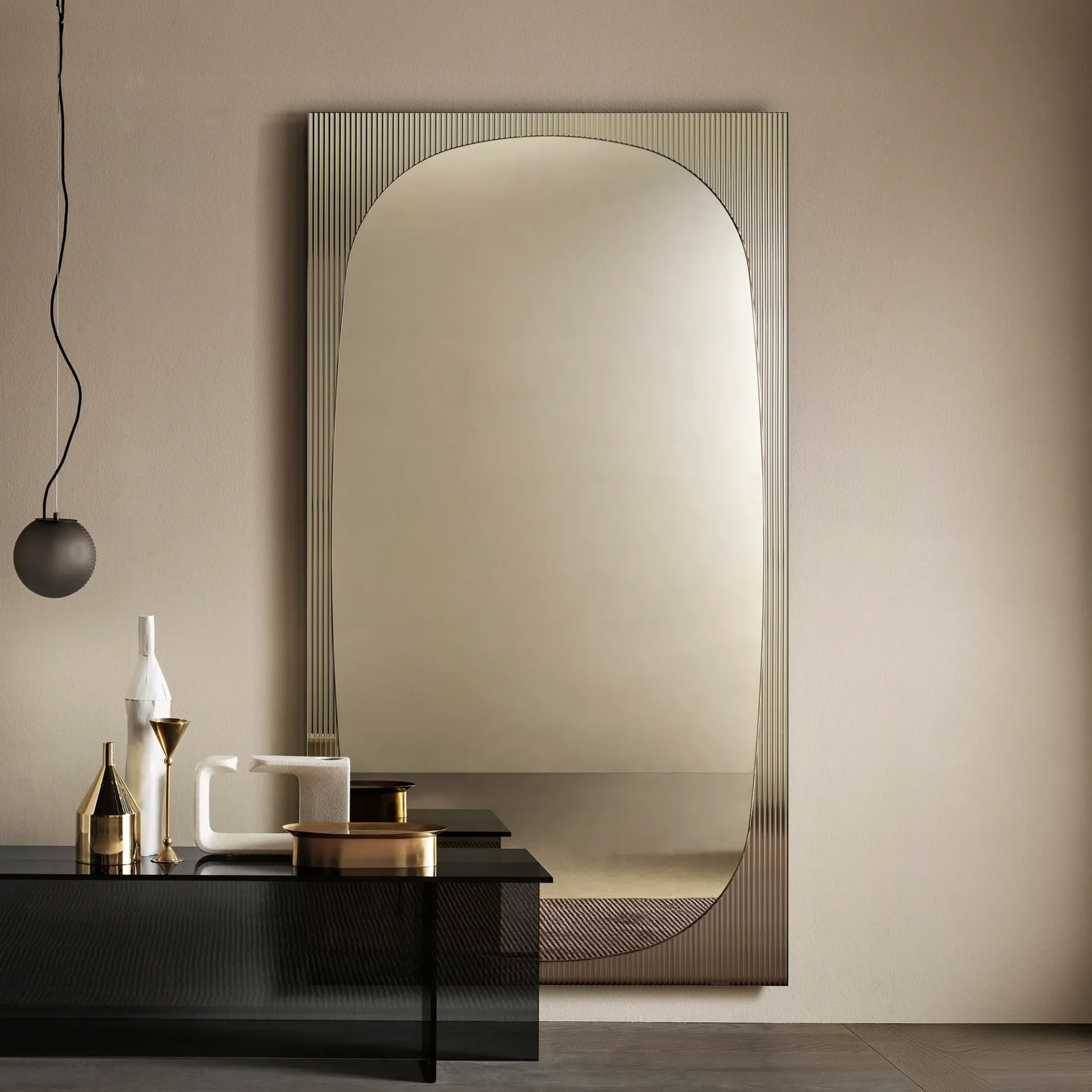 A modern interior with a large, arched mirror with a ribbed frame above a dark console table adorned with white and gold decorative objects. A dark pendant light hangs on the left.