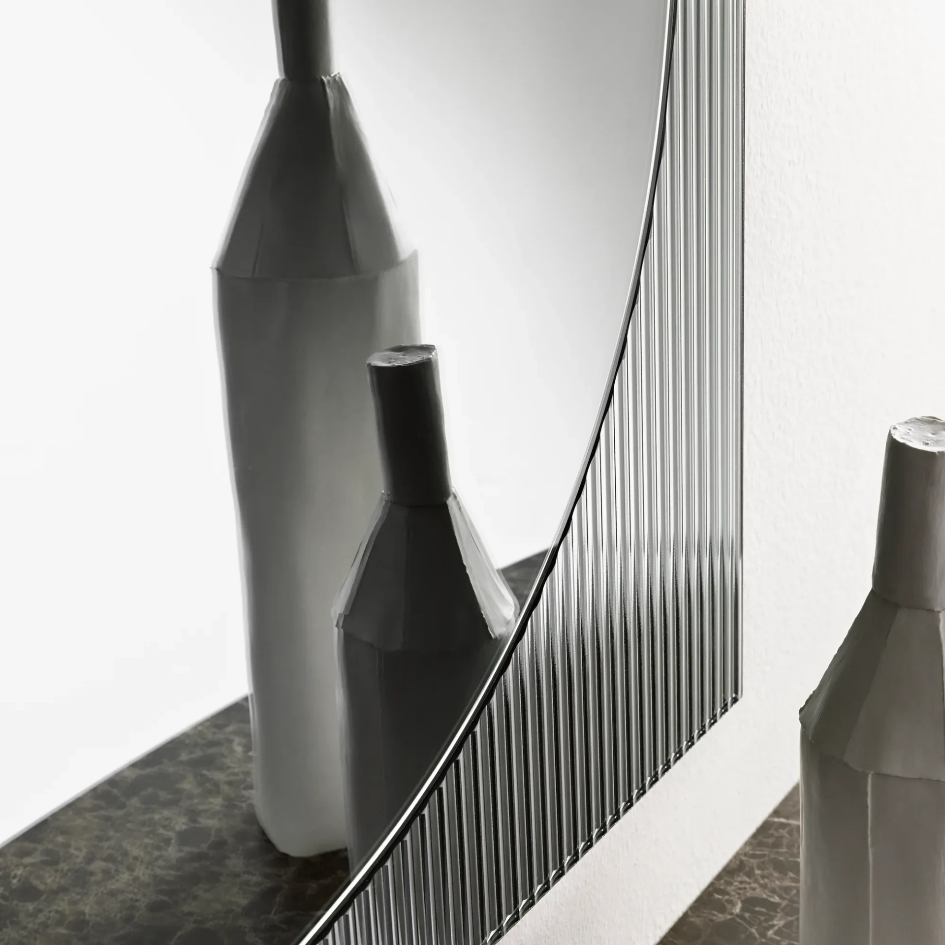 Three light grey faceted vases, one reflected, on a dark marble surface next to a curved mirror with a fluted frame, against a white wall.