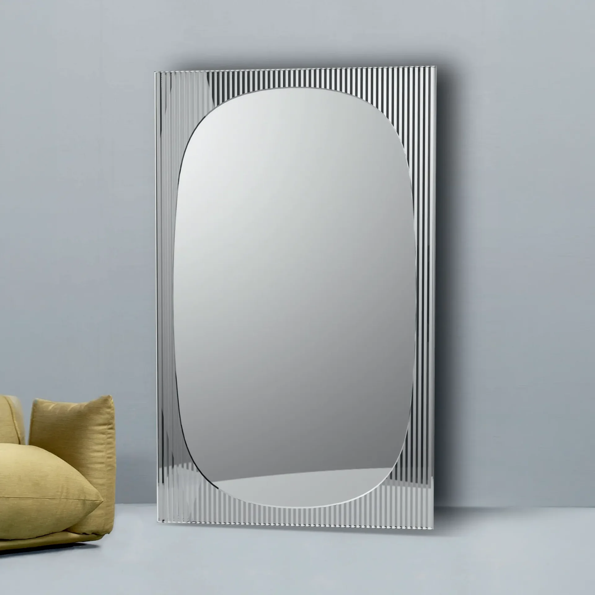 A tall, rectangular mirror with an oval reflective surface and a fluted, striped frame stands against a light gray wall. Part of a yellow armchair is visible on the left.
