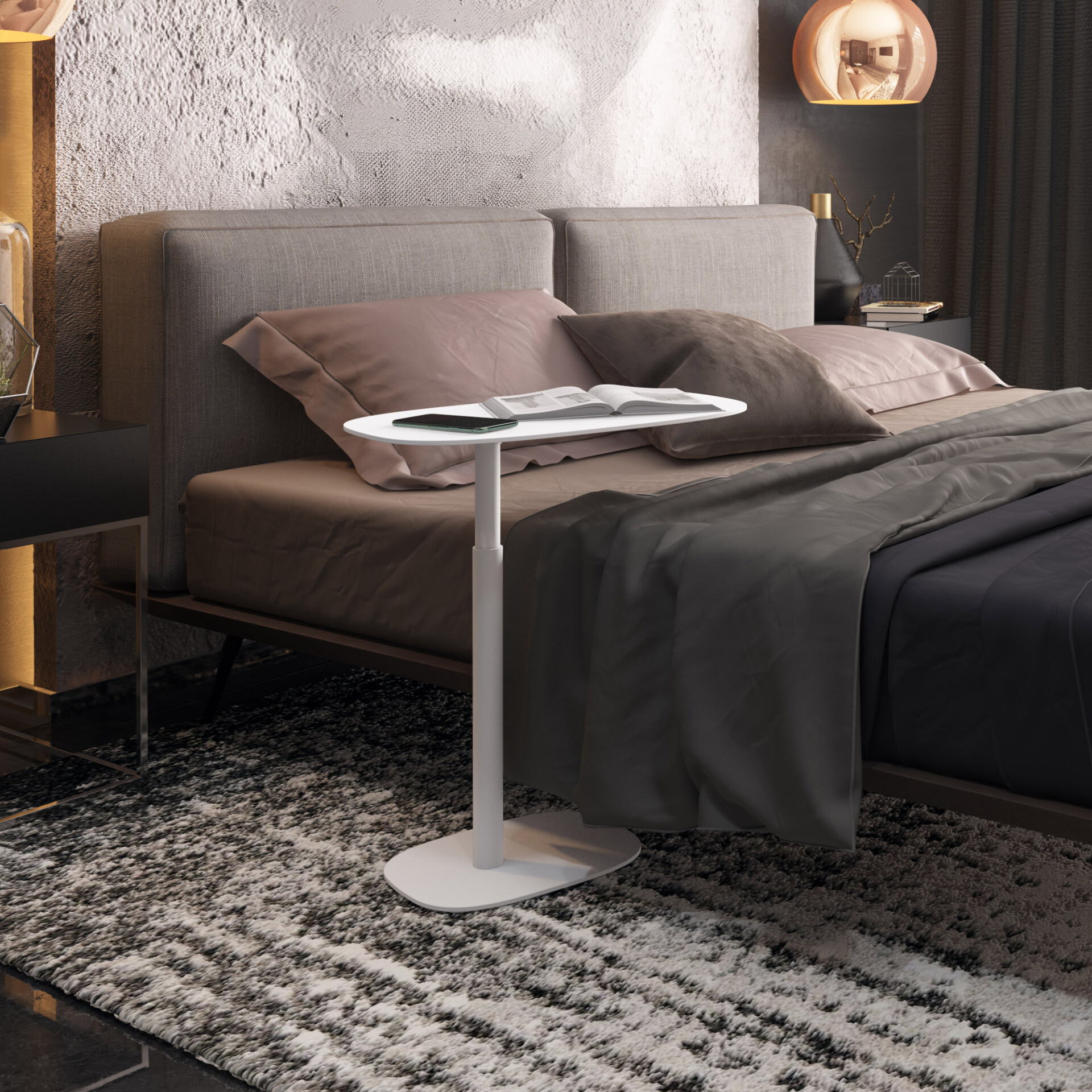 A modern bedroom with a gray upholstered bed and dark gray bedding. A white, adjustable over-bed table with a book and phone is next to the bed. The room features textured walls, warm lighting, and a shaggy gray rug.