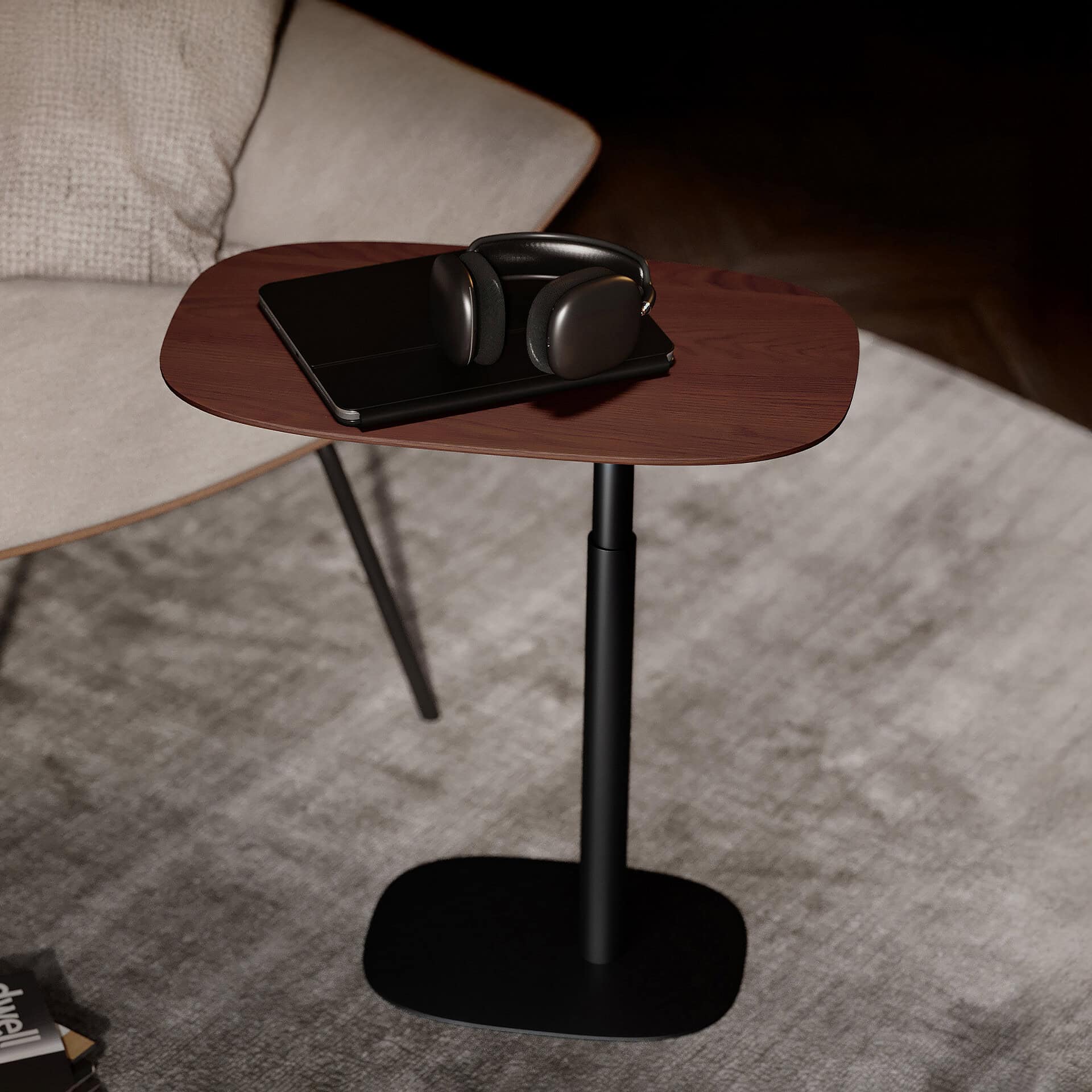 A modern, adjustable side table with a dark wood top, black stand, and base. A black tablet and dark grey over-ear headphones rest on the table. A beige armchair and a grey rug are in the background.
