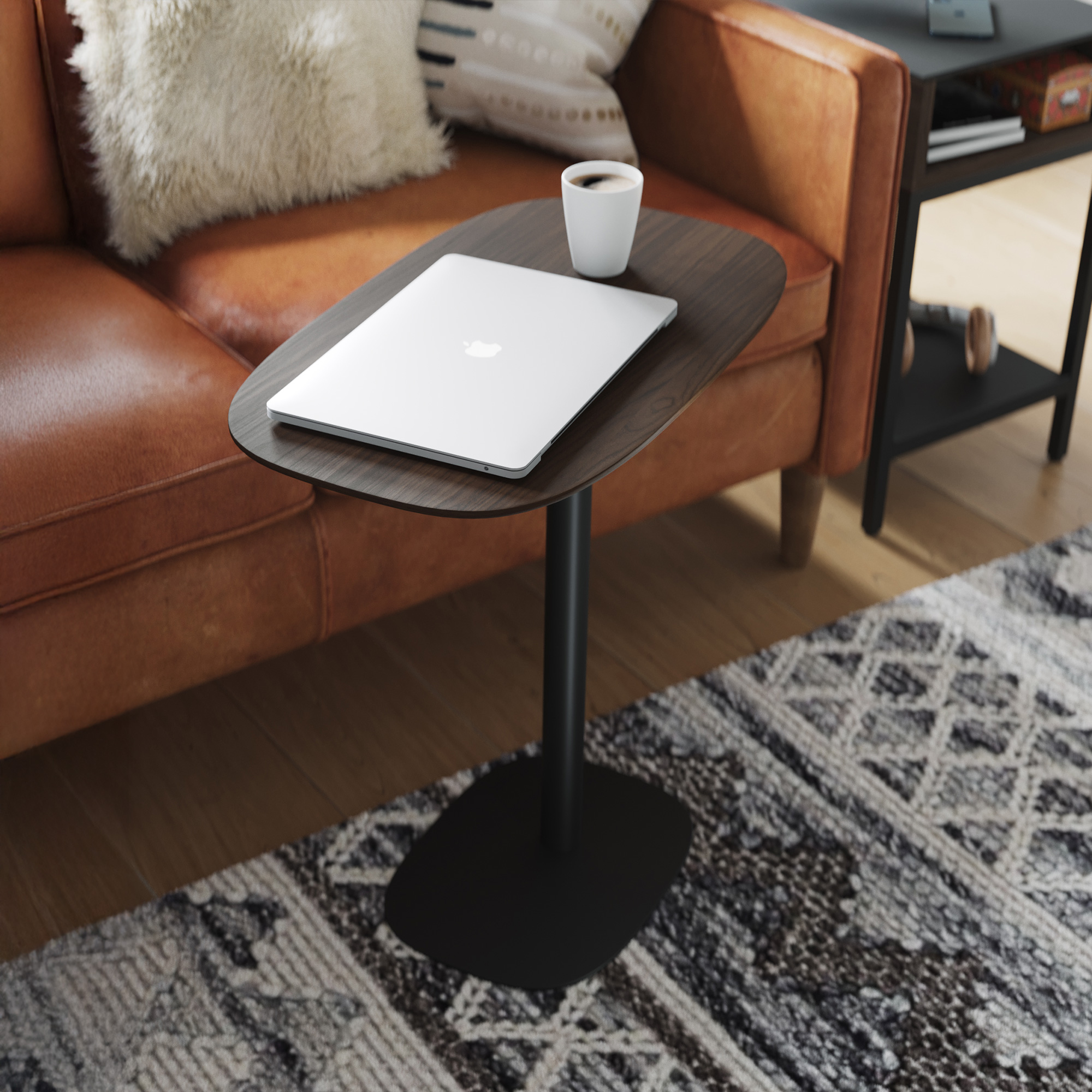 A silver laptop and a white coffee mug on a dark wood C-table next to a brown leather sofa with a faux fur pillow.