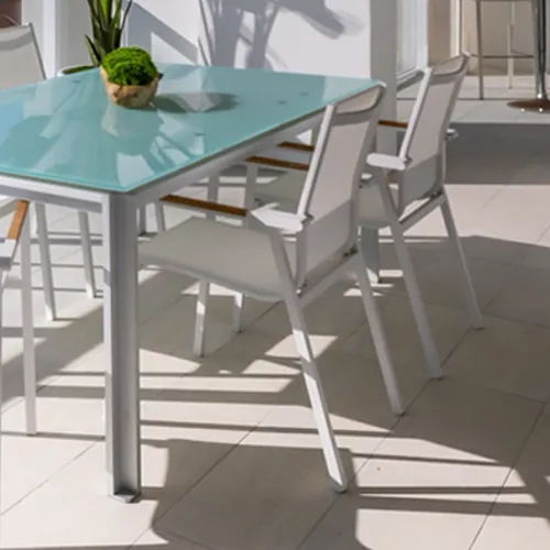 Outdoor dining set with a white metal table and light blue glass top, two white chairs with wooden armrests, and a small green plant on the table, all casting long shadows on a tiled patio.