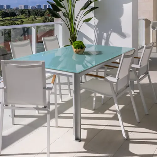 A modern outdoor dining set with a frosted glass table and white chairs on a balcony, overlooking a golf course and city skyline.