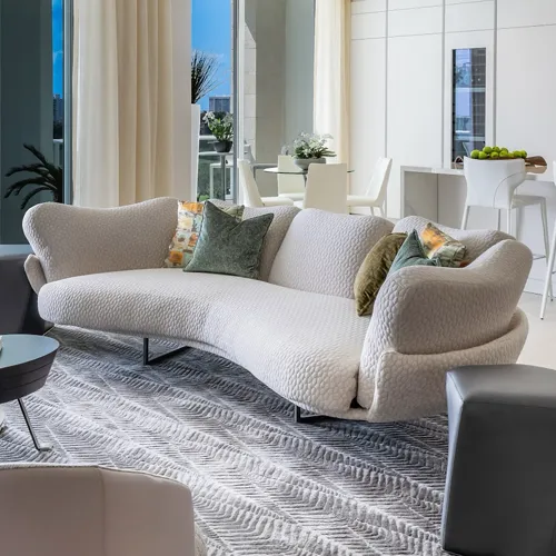 A modern living room with a large, light-colored curved sofa, patterned rug, and a dining area visible in the background.
