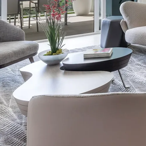 A modern living room with a two-tiered, organic-shaped coffee table, a plant with red flowers, a book, and various armchairs on a patterned rug.