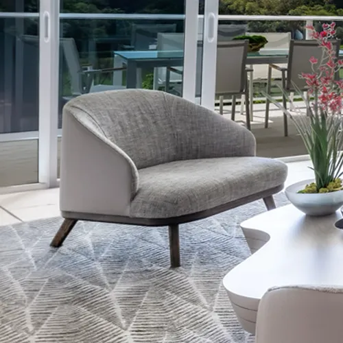 A gray loveseat with a textured fabric and light gray sides sits on a geometric patterned rug. Through glass doors in the background, an outdoor patio with a dining table and chairs is visible, surrounded by green foliage. A white coffee table with a floral arrangement is in the foreground.