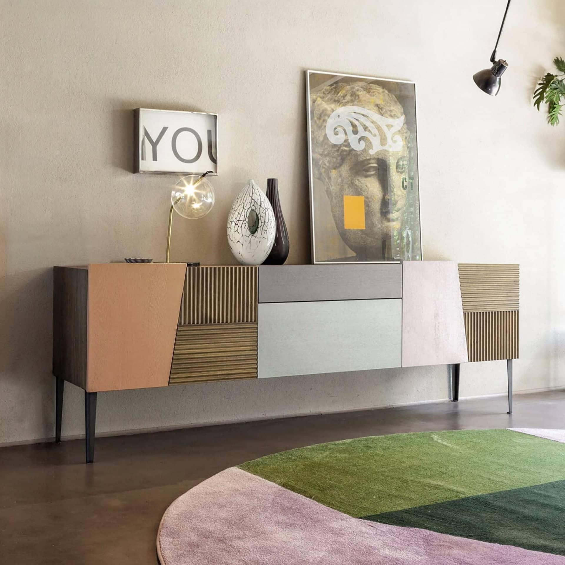 A modern living room featuring a long, multi-colored geometric sideboard, a round rug with green and pink sections, and contemporary wall art including a classical bust print and a 'YOU' sign.
