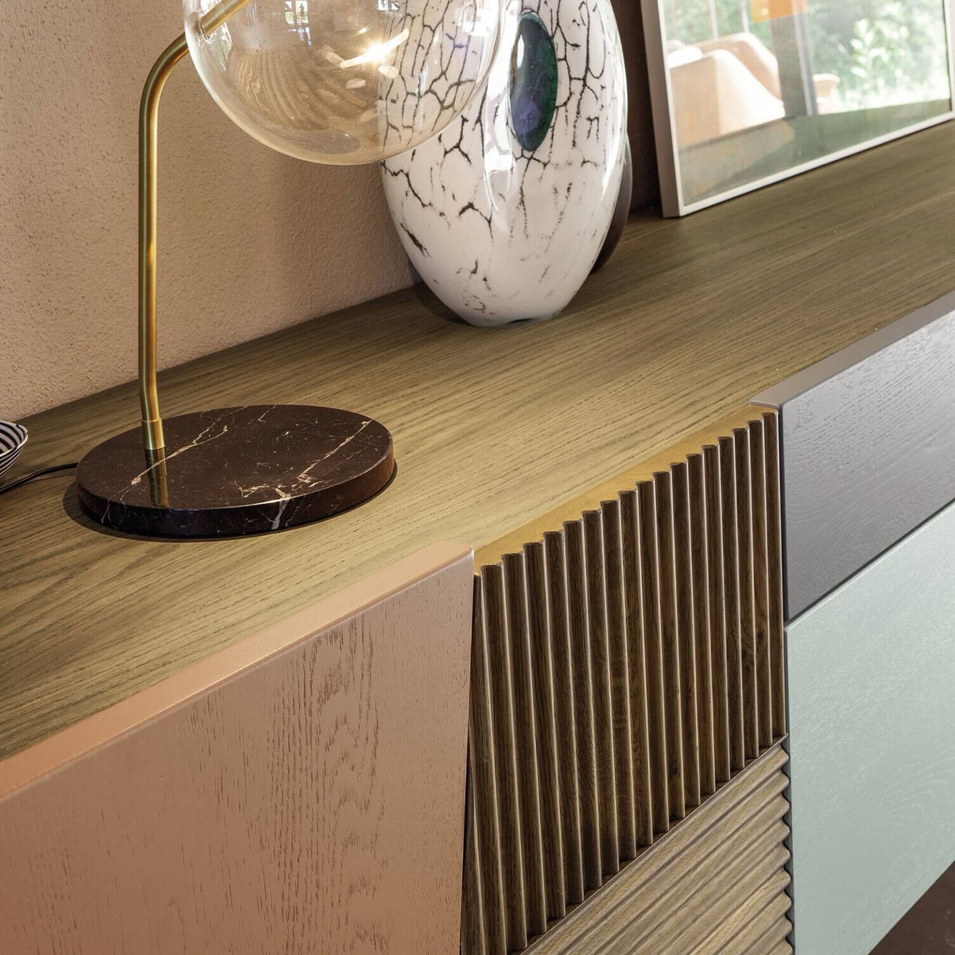 Close-up of a modern sideboard with a brass lamp on a marble base, a white crackle-glazed vase, and multi-colored cabinet panels featuring fluted wood and matte finishes in brown, green, and grey.