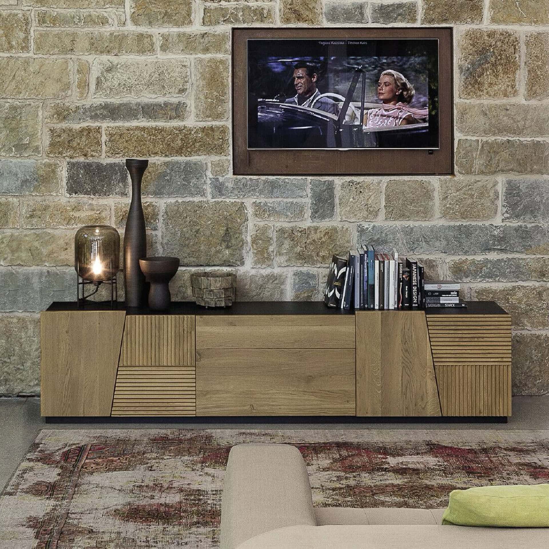A modern wooden media console with a dark top sits against a rustic stone wall. A TV above shows a movie scene. A lamp, vases, and books decorate the console. A patterned rug and part of a light-colored sofa with a green pillow are in the foreground.