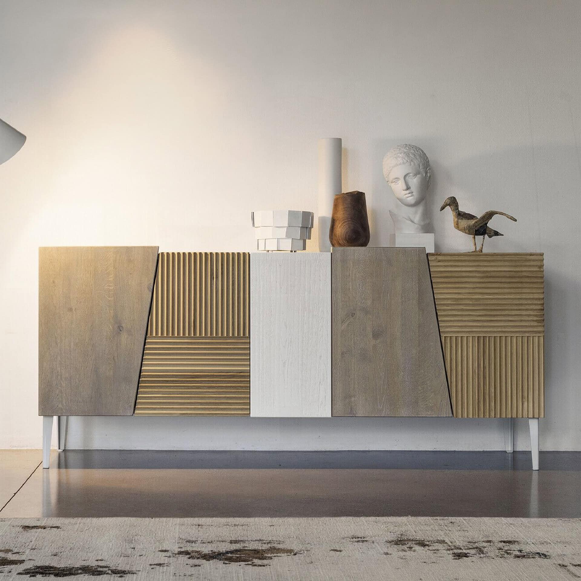 A modern sideboard with a multi-textured facade in light grey wood, white, and golden fluted panels, styled with decorative objects on top, including a classical bust and a bird sculpture.
