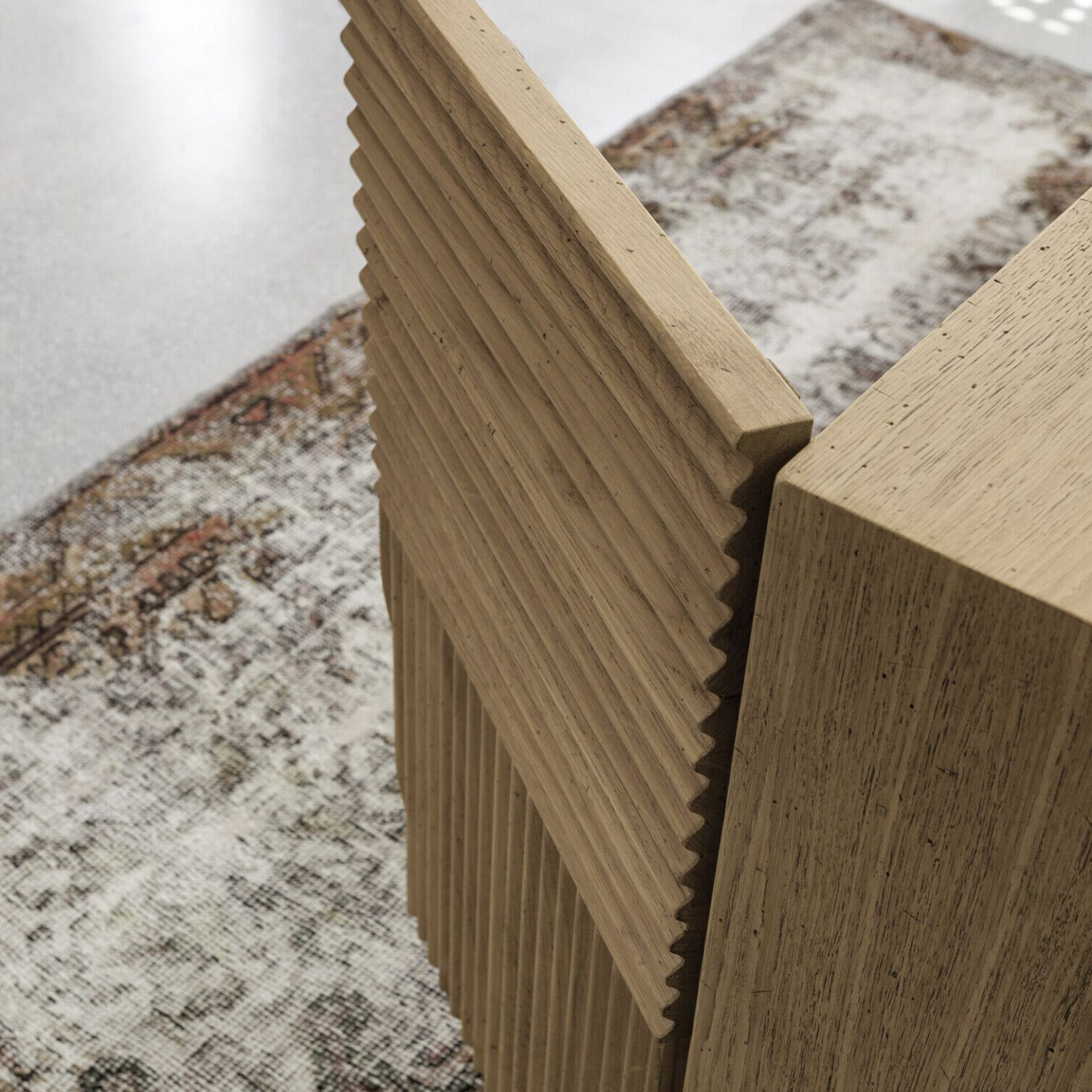 Close-up of light fluted wood furniture with visible grain, next to a block of plain wood, resting on a faded patterned rug.