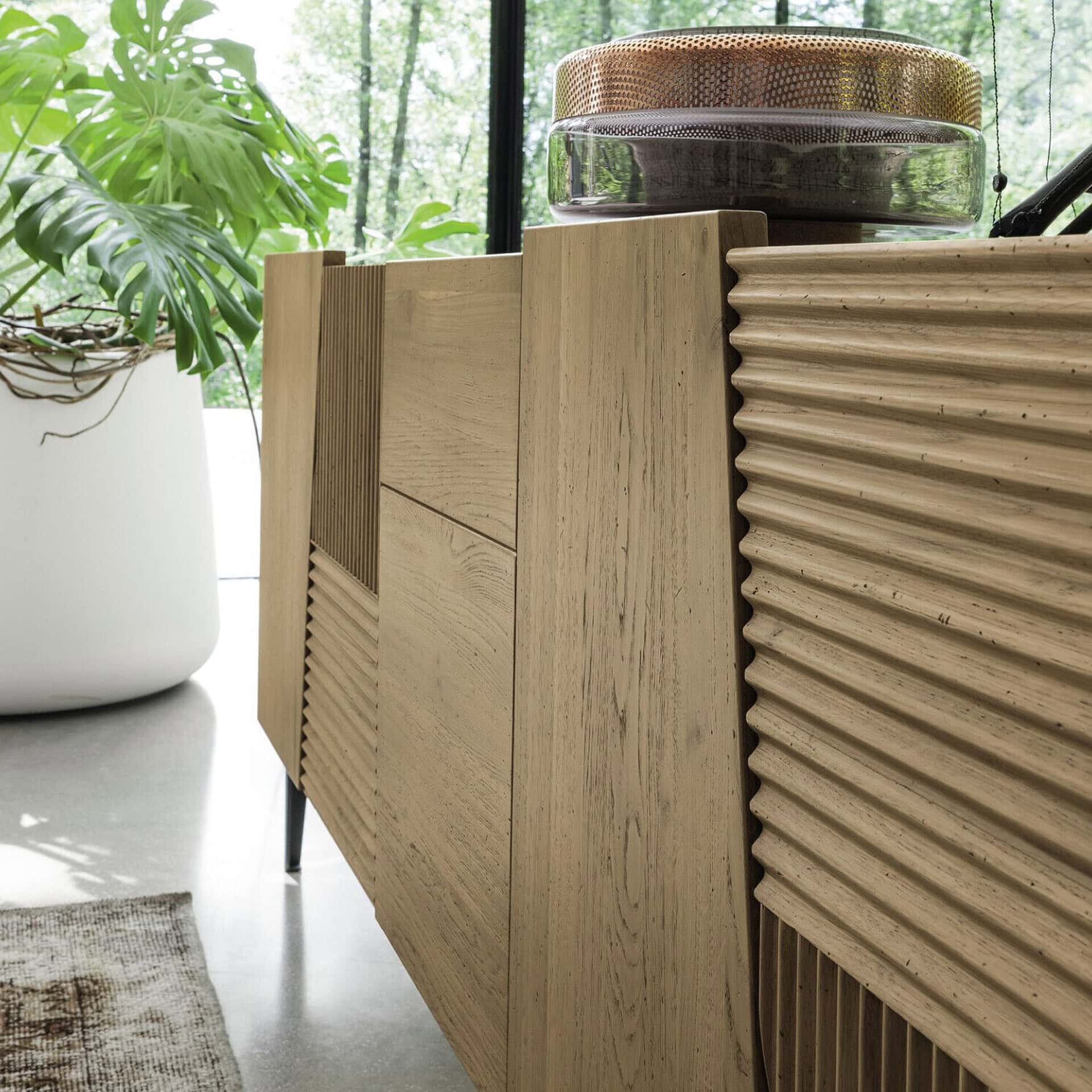 A modern light wood sideboard with textured geometric panels, a large Monstera plant in a white pot, and a decorative glass and perforated metal object on top, set against a window overlooking green trees.
